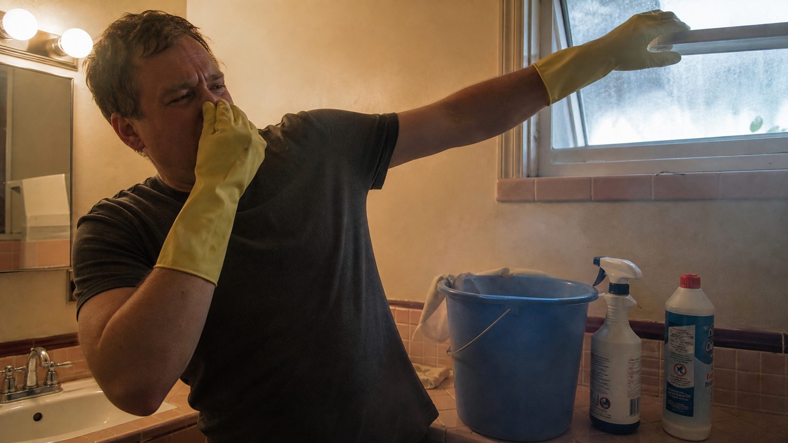A person in a home bathroom recoiling and covering their nose and mouth while reaching to open a window, a cleaning bucket with two bottles beside it on the counter, hazy air visible in the light