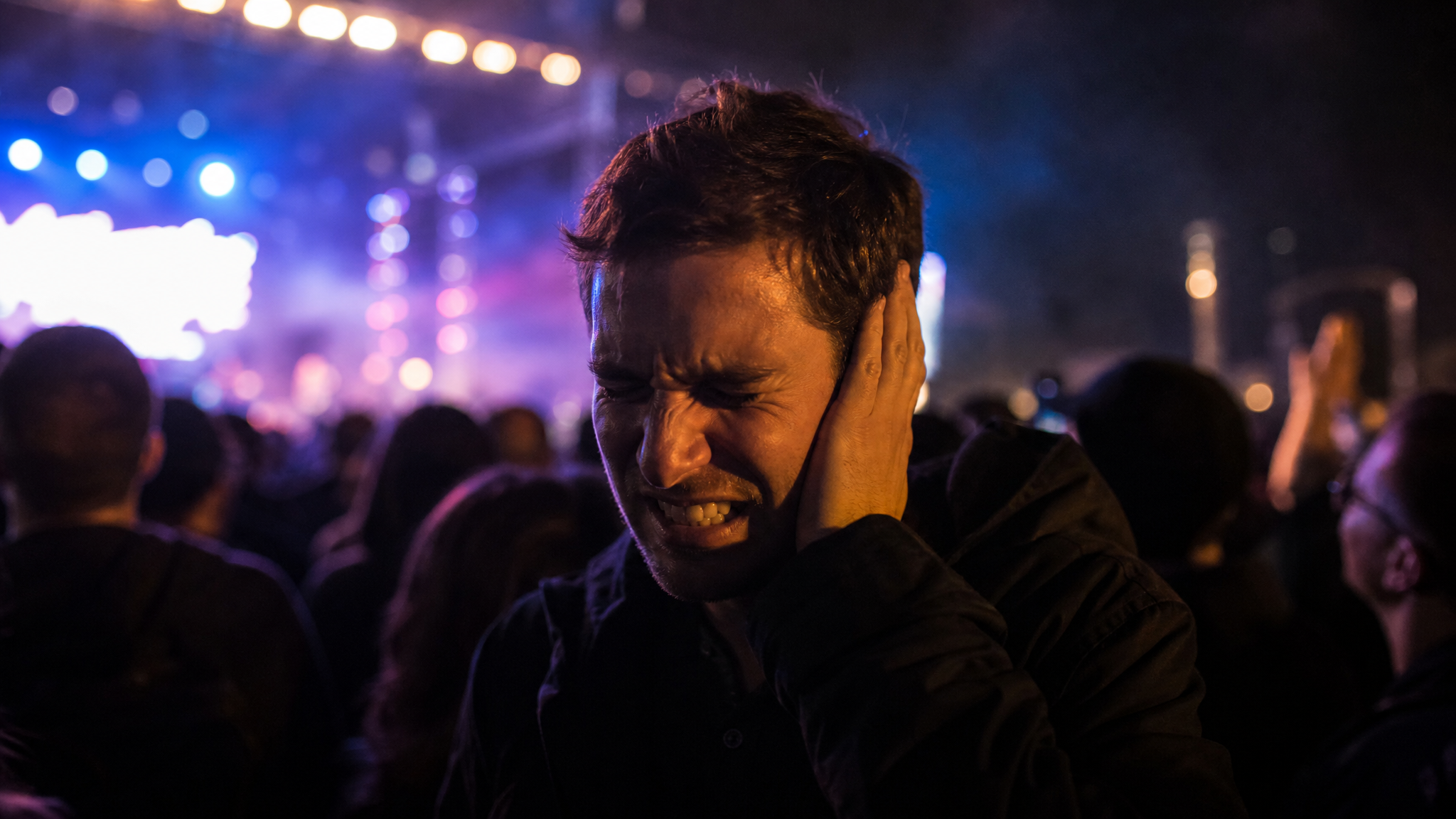 A man in his 30s at a loud concert at night grips the side of his head, wincing in pain, colored stage lights glowing behind him