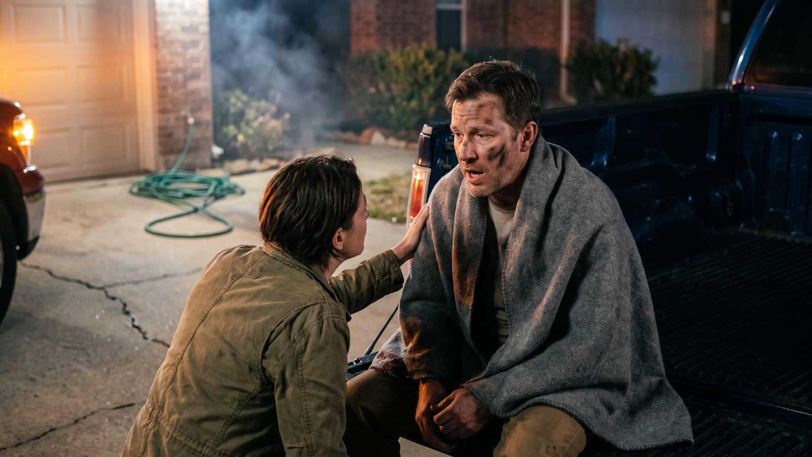 A man in his forties sits on a tailgate in a driveway at night wrapped in a blanket, face streaked with soot, while another adult crouches beside him listening to him speak, smoke haze and emergency light glow behind them