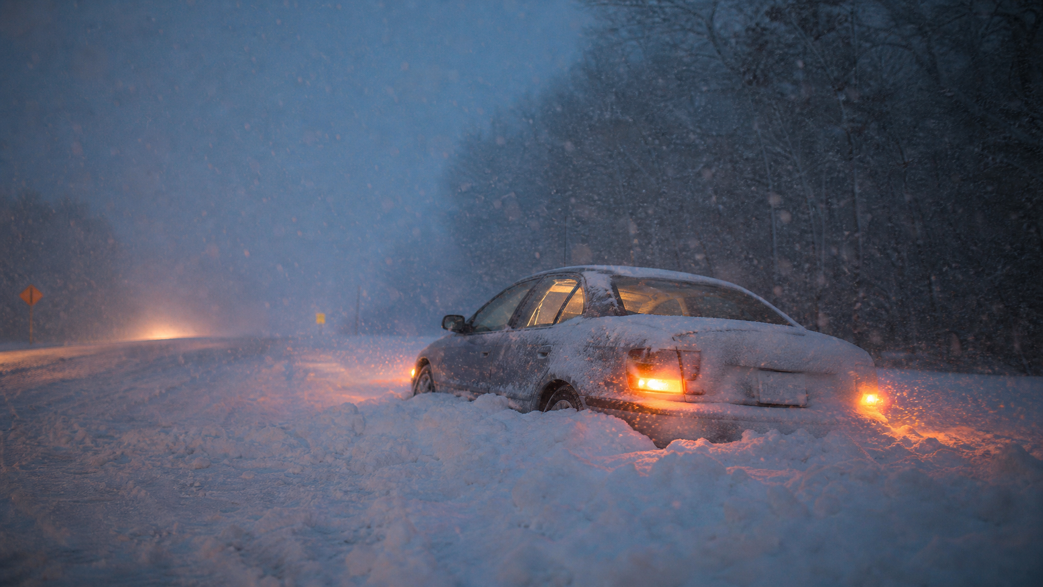 A car sits stranded in deep snow on a rural road at dusk with hazard lights glowing through heavy falling snow