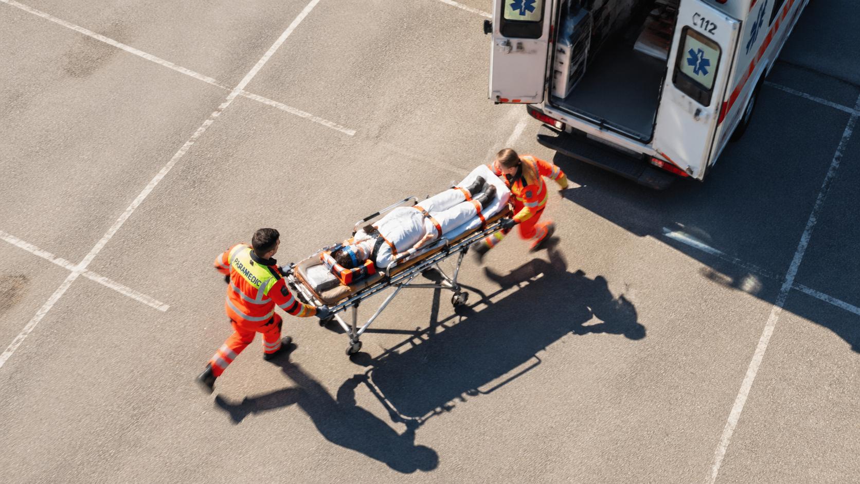 Aerial view of a two-person EMT team rushing with a stretcher across a sunlit parking lot toward an ambulance with open rear doors, dynamic motion, photojournalistic documentary style