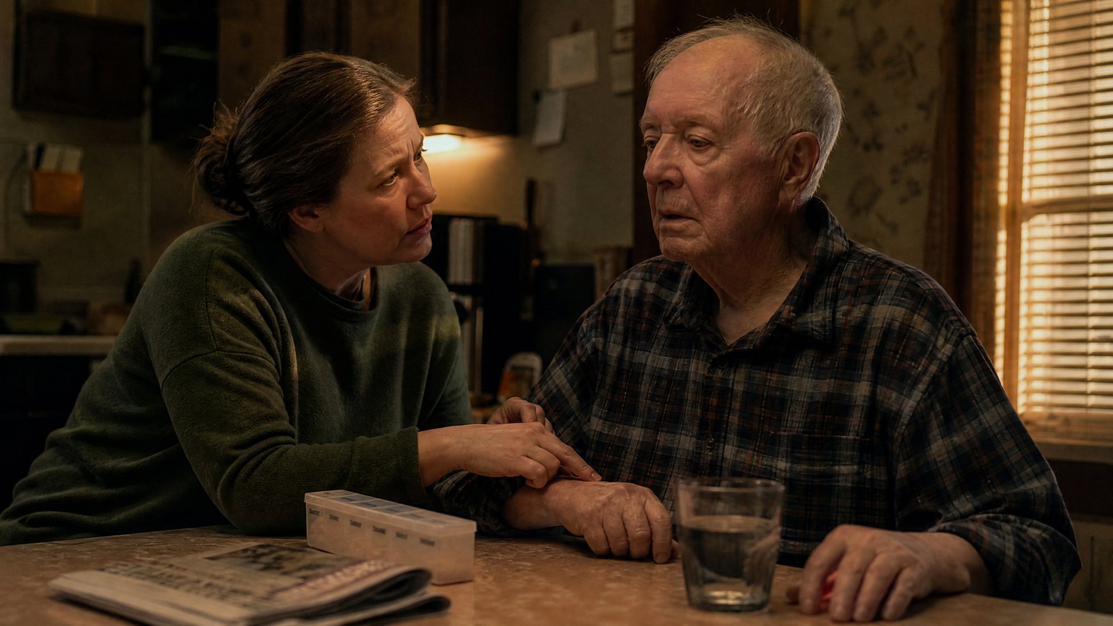 An adult daughter crouches beside her elderly father in a dim kitchen, one hand on his wrist taking a pulse while he looks vacantly past her