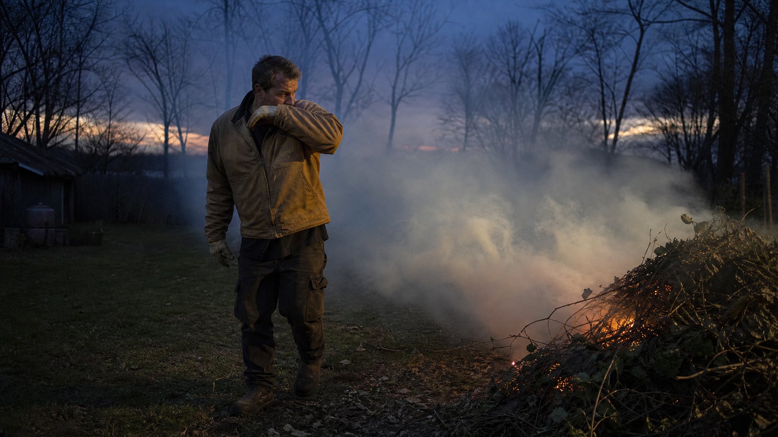 An adult man in a work jacket stepping back from a smoldering brush pile at dusk as thick smoke drifts across a rural yard