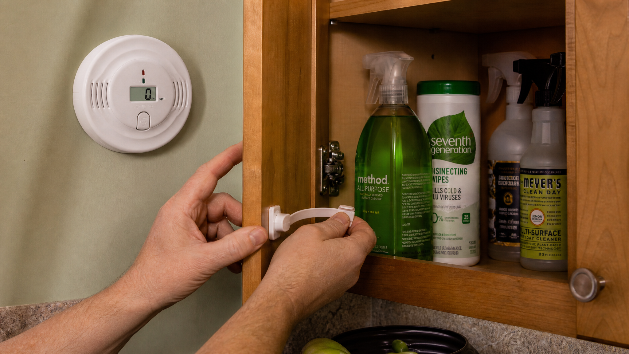 An adult's hands installing a childproof latch on a lower kitchen cabinet stocked with cleaning products in their original labeled bottles, a carbon monoxide alarm visible on the wall nearby, warm home lighting