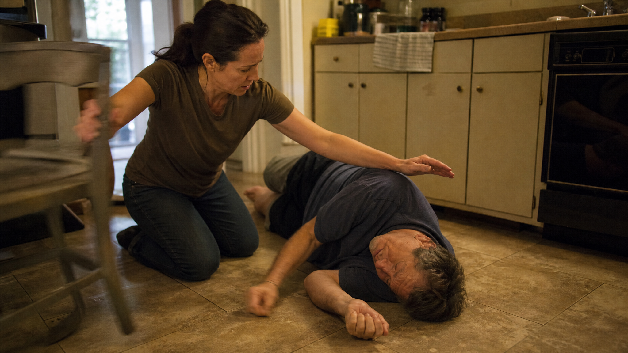 Adult kneeling on a kitchen floor beside a person having a seizure, clearing a chair out of the fall path