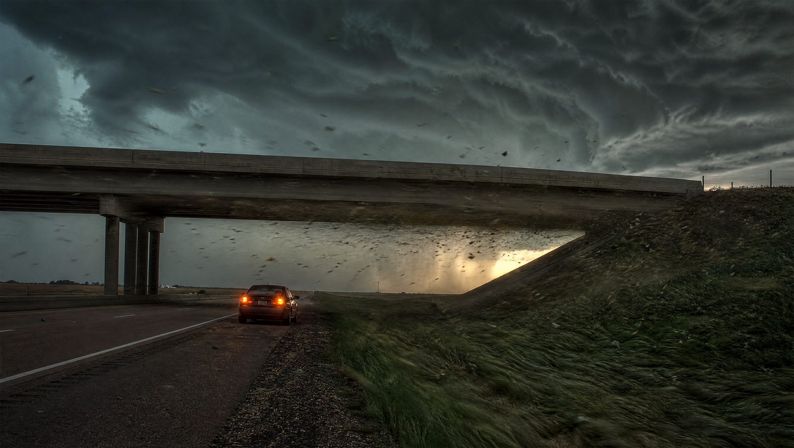 A highway overpass during a dark storm with wind and debris funneling through the narrow space beneath it, showing why it is not a safe tornado shelter