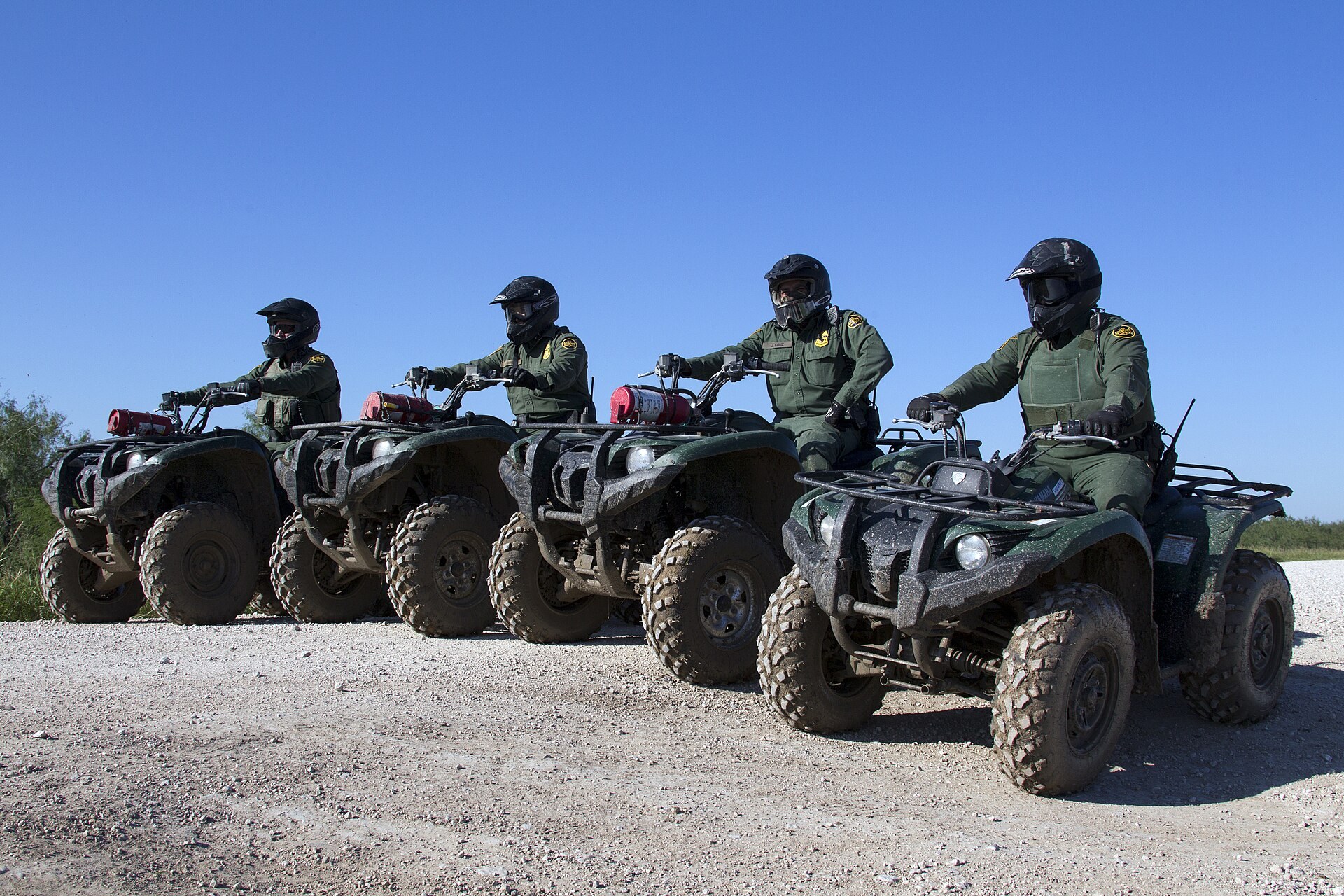Four Border Patrol agents in green uniforms and helmets sitting on all-terrain vehicles along a gravel road in South Texas