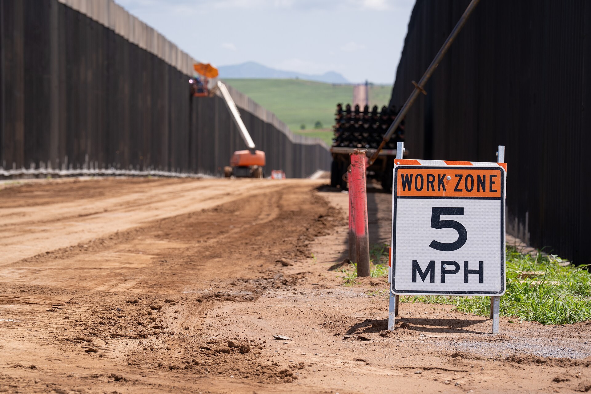 Tall dark steel border wall panels on both sides of a dirt road with a work zone sign and a lift truck near Lochiel, Arizona