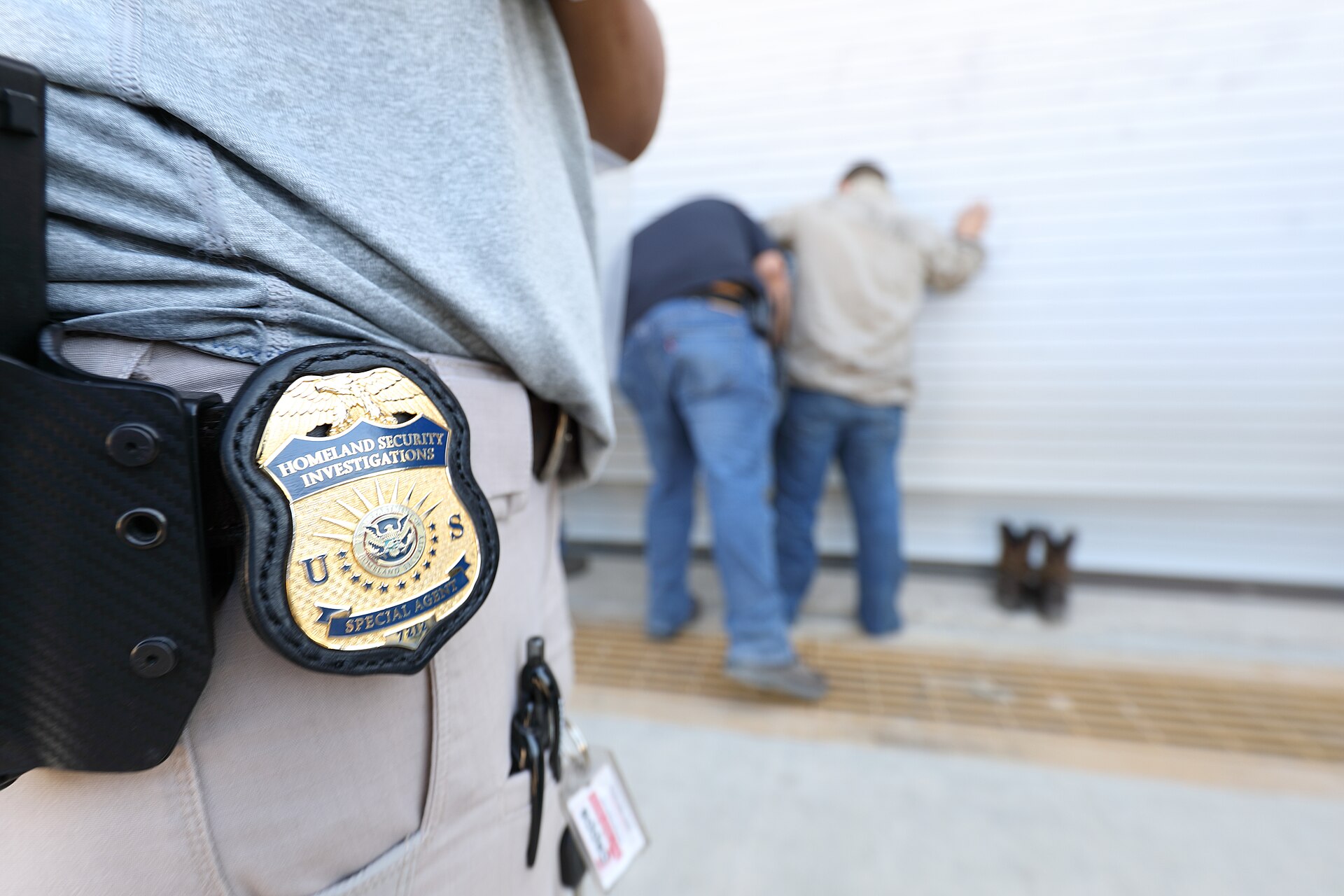 Homeland Security Investigations special agent badge in the foreground as agents detain a person against a wall during a search warrant operation