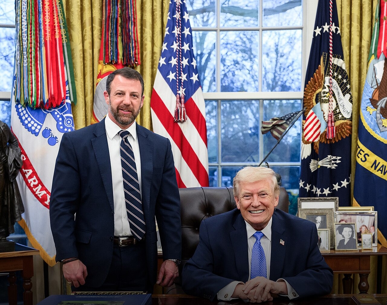 DHS Secretary Markwayne Mullin standing beside President Trump seated at the Resolute Desk in the Oval Office