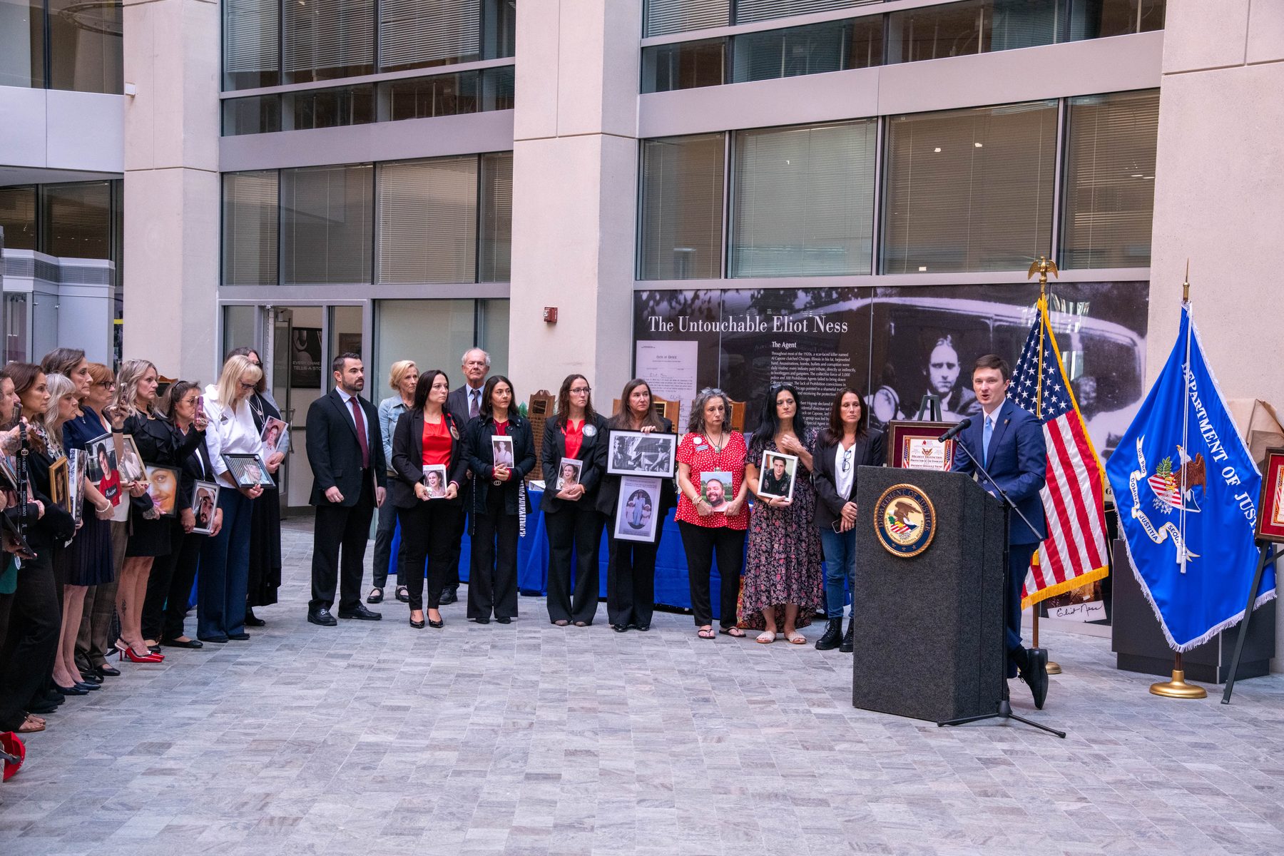 Family members holding framed photos of loved ones stand in a courtyard as an official speaks at a Justice Department podium flanked by American and DOJ flags