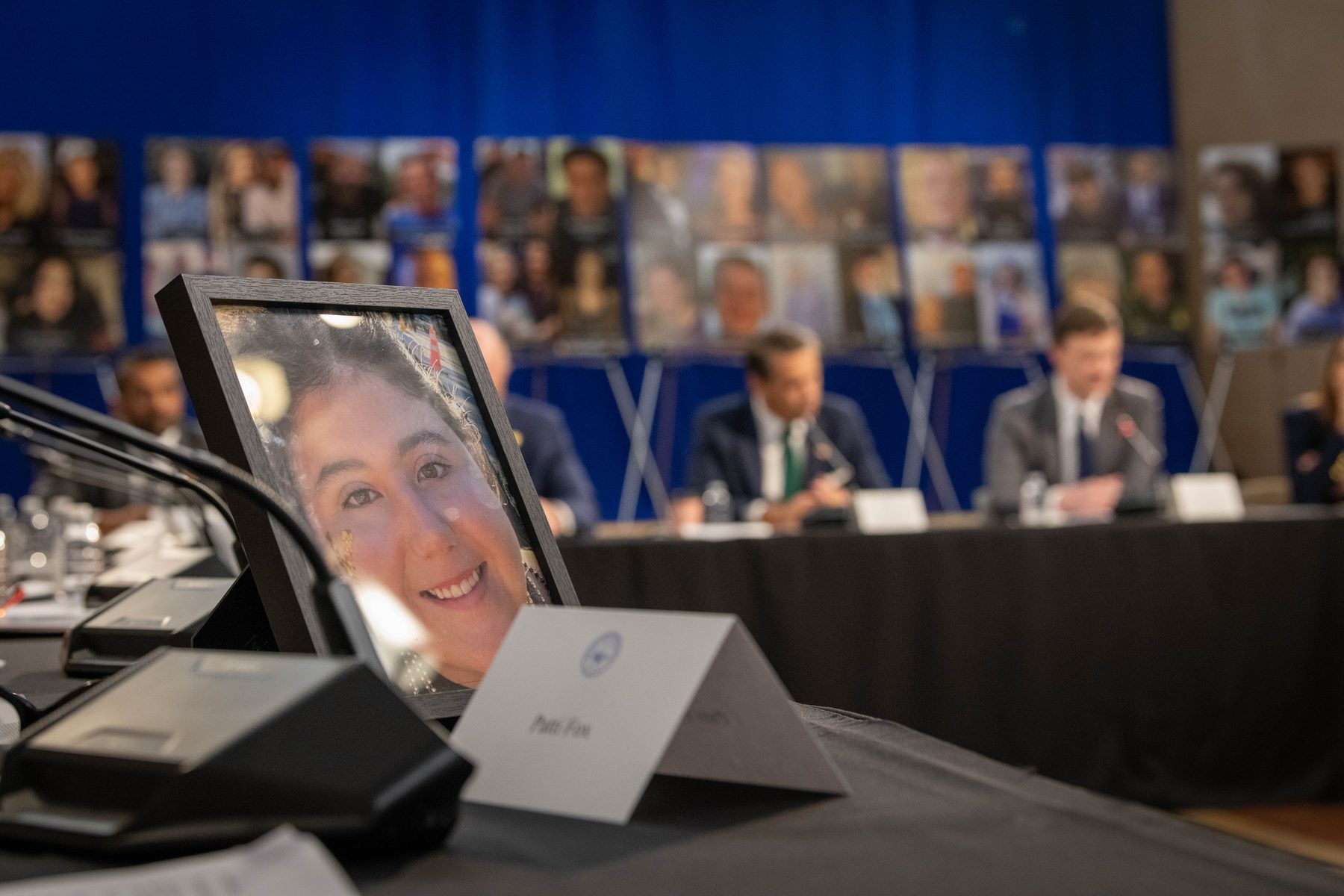 A framed photo of a smiling young woman on the summit table at the seat marked Patti Fox, with the panel and victim photos blurred behind