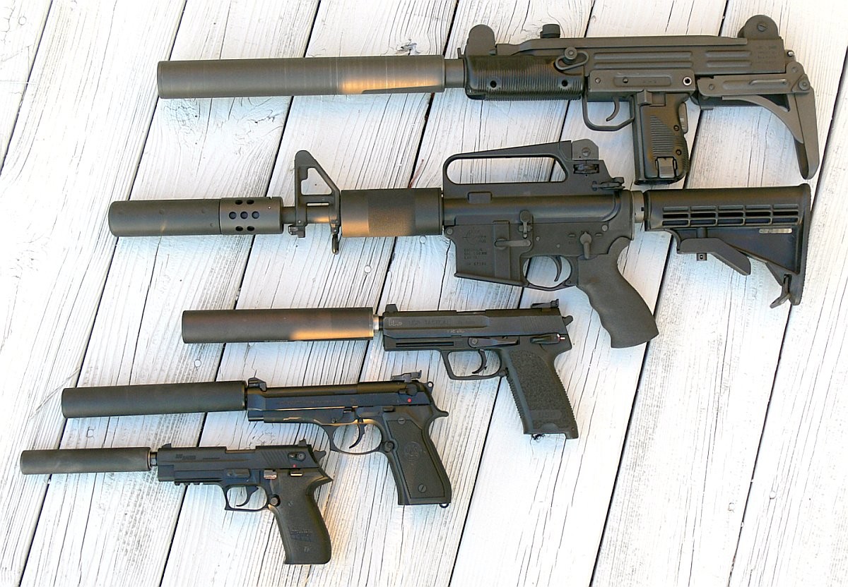 Five suppressed firearms laid out on white boards, including a suppressed Uzi, an AR-15, an HK USP Tactical, a Beretta 92FS and a SIG pistol