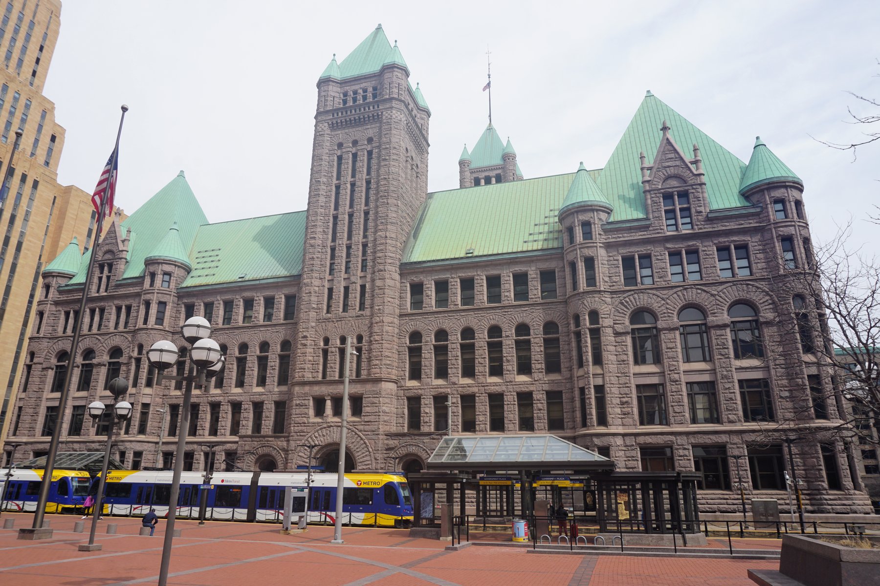 Minneapolis City Hall, a red granite Romanesque building with green copper roofs, with a light rail train passing in front