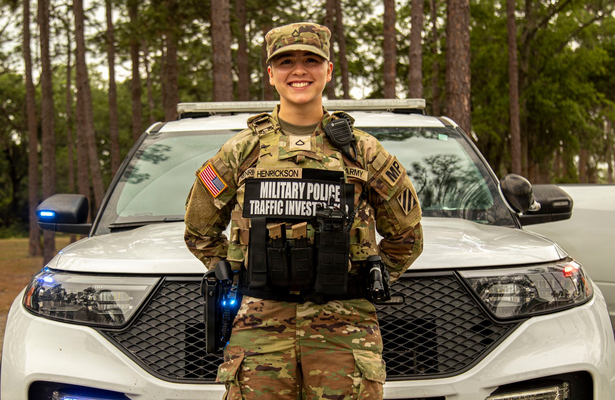 A soldier in a modern tactical vest with a Military Police patch standing in front of a patrol vehicle