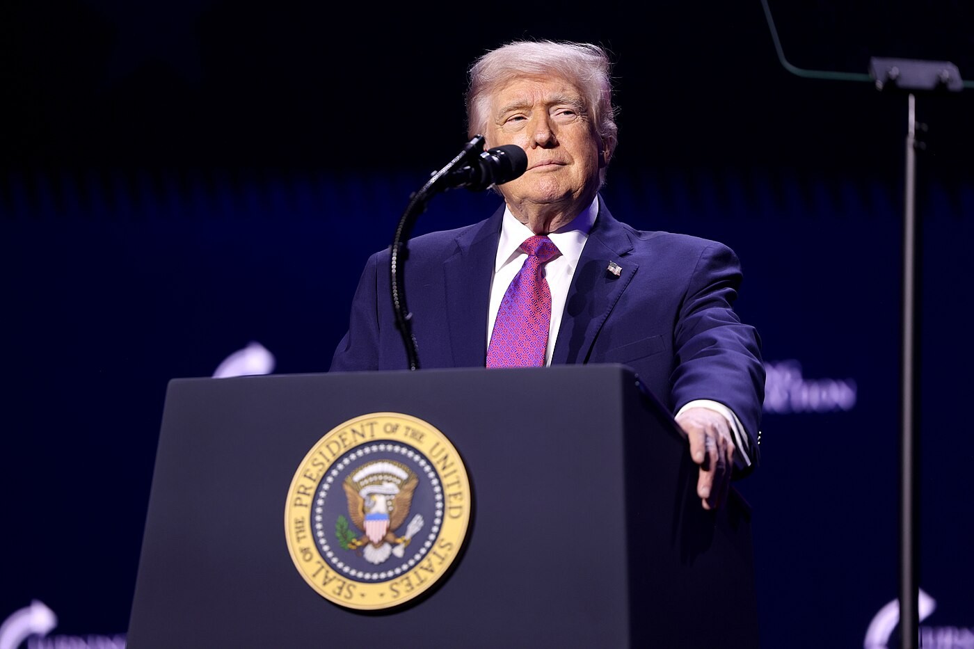 President Donald Trump speaking at a podium with the presidential seal at a Turning Point Action rally