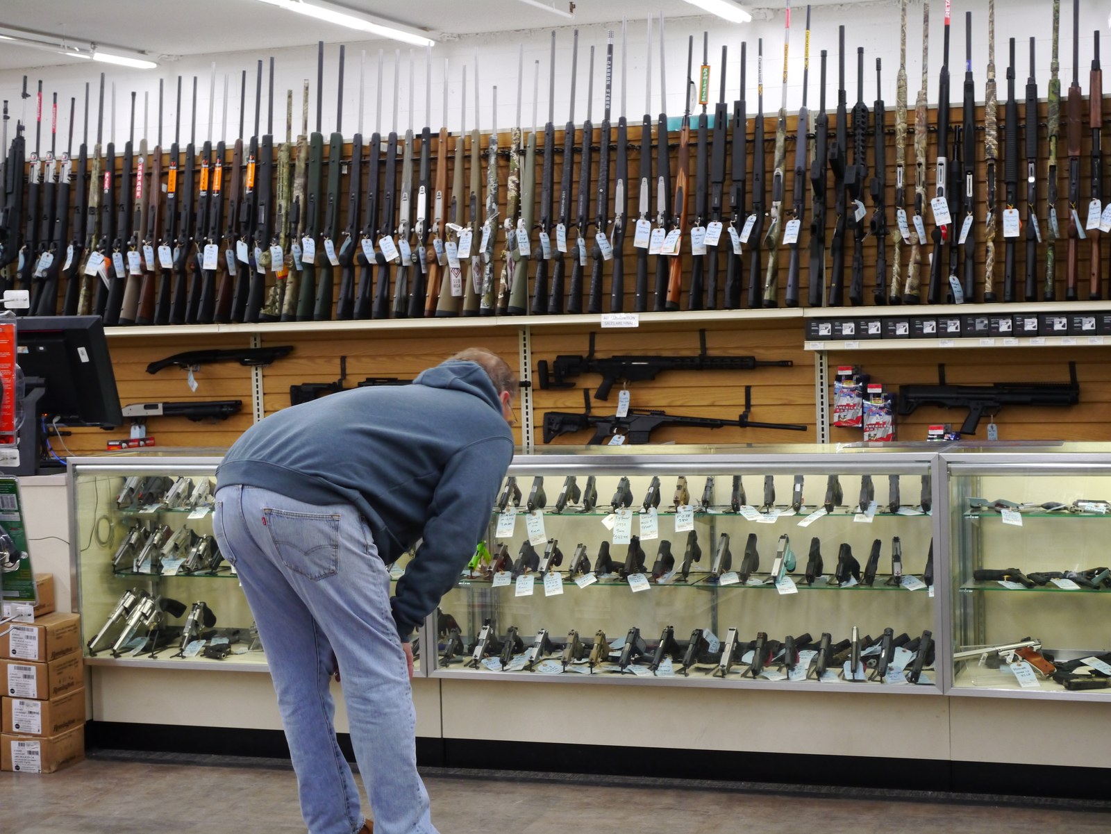 Handguns displayed in a glass case at a licensed gun store while a customer looks over the selection