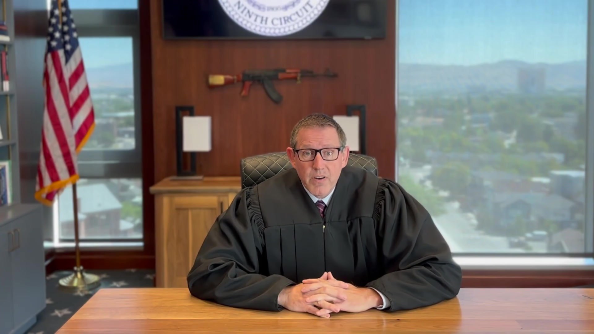 Ninth Circuit Judge Lawrence VanDyke seated at a desk in judicial robes, with a rifle mounted on the wall behind him