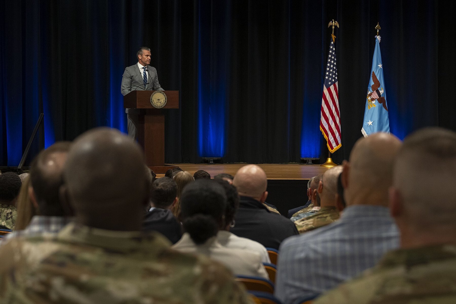 Service members and civilians in an auditorium facing a lectern flanked by flags during the Secretary of War's prayer service at the Pentagon