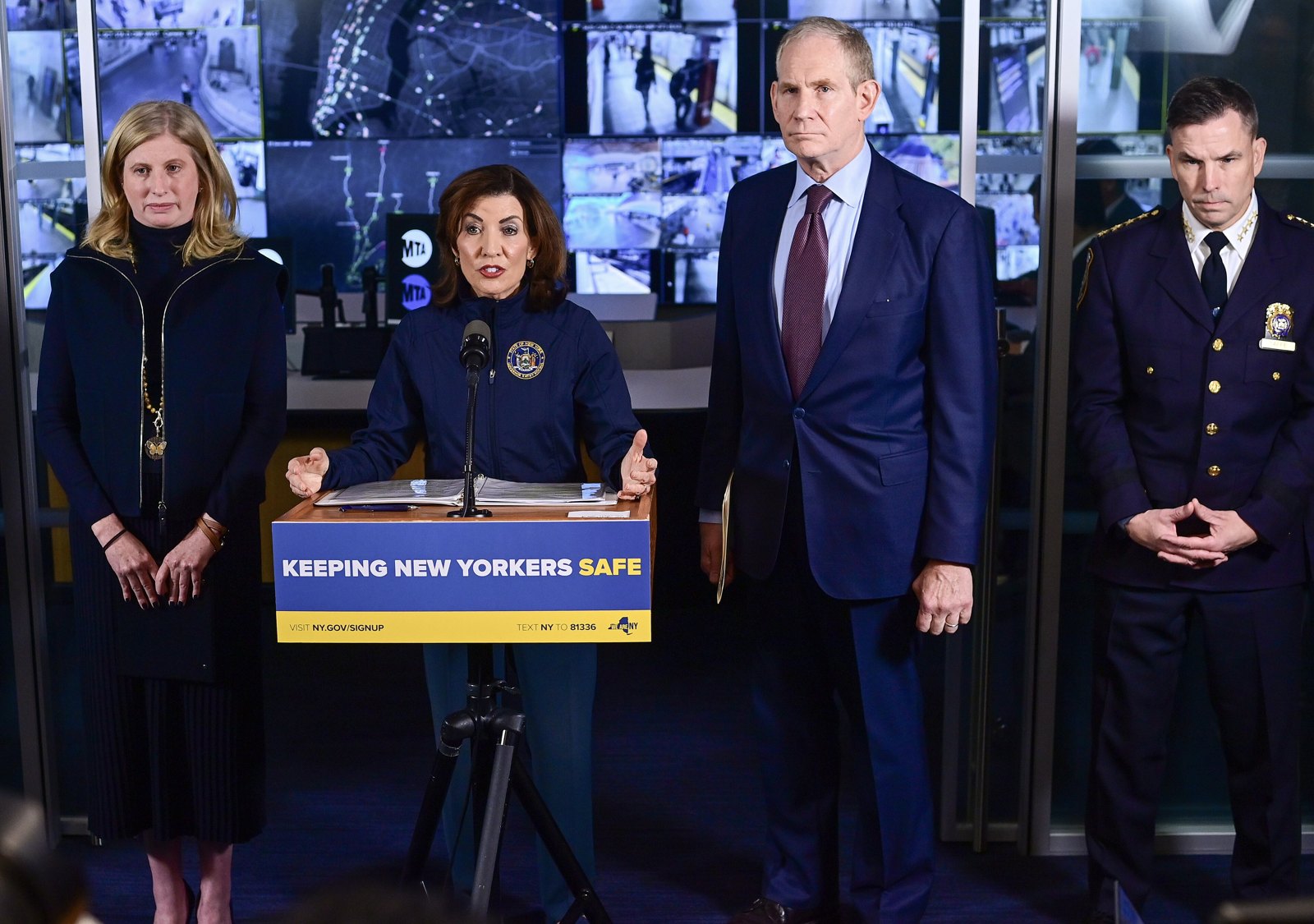 Gov. Kathy Hochul speaking at a podium reading Keeping New Yorkers Safe, with officials standing beside her