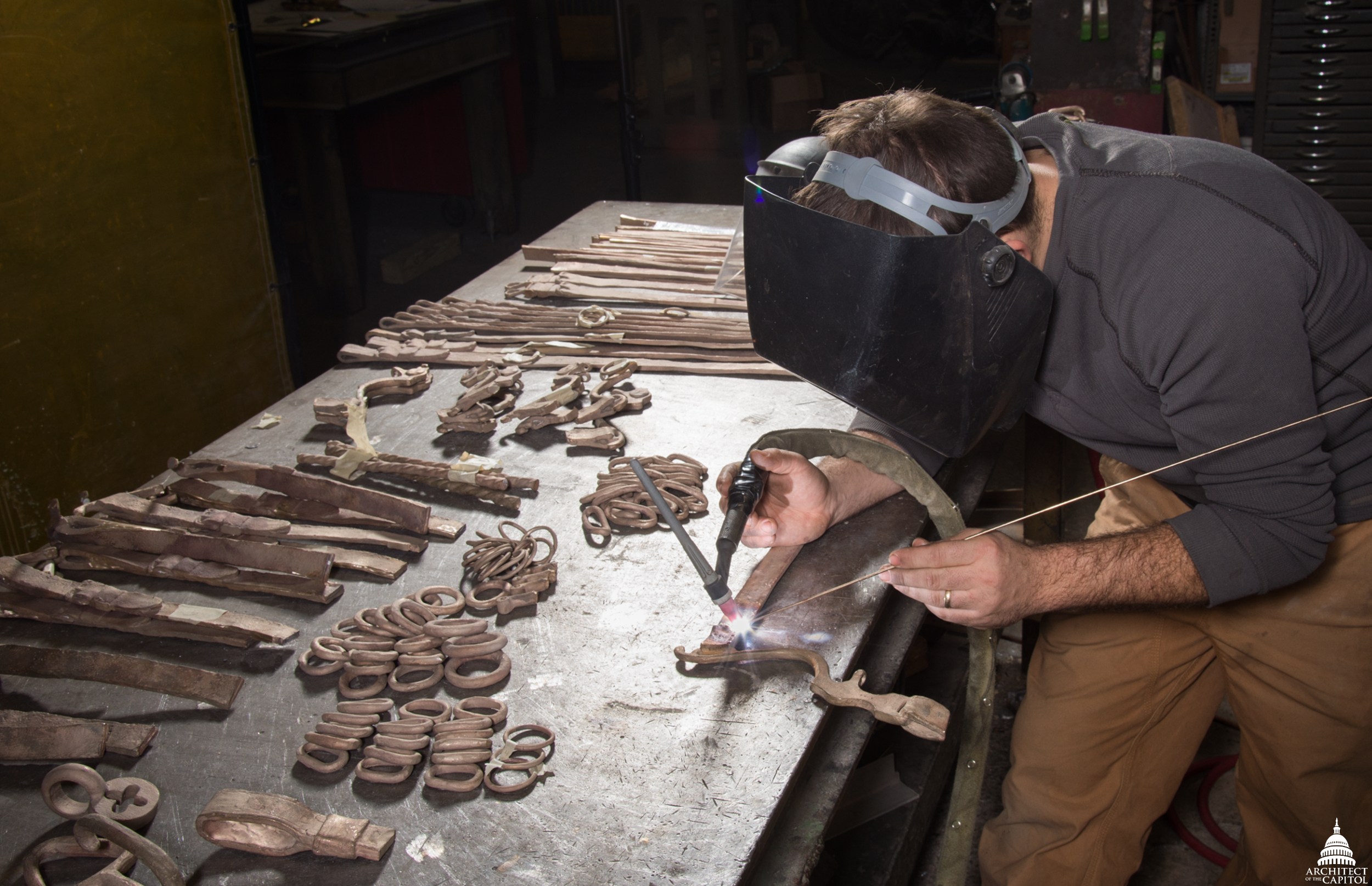A metalworker in a welding hood torches cast bronze elements laid out on a workbench at a Philadelphia foundry