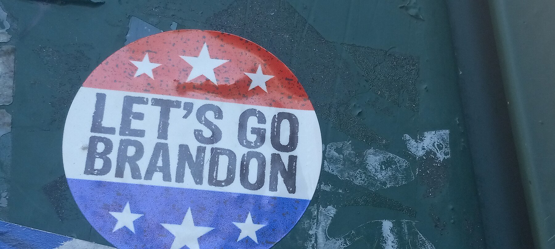 Red, white and blue round sticker reading Let's Go Brandon stuck to a dark green surface