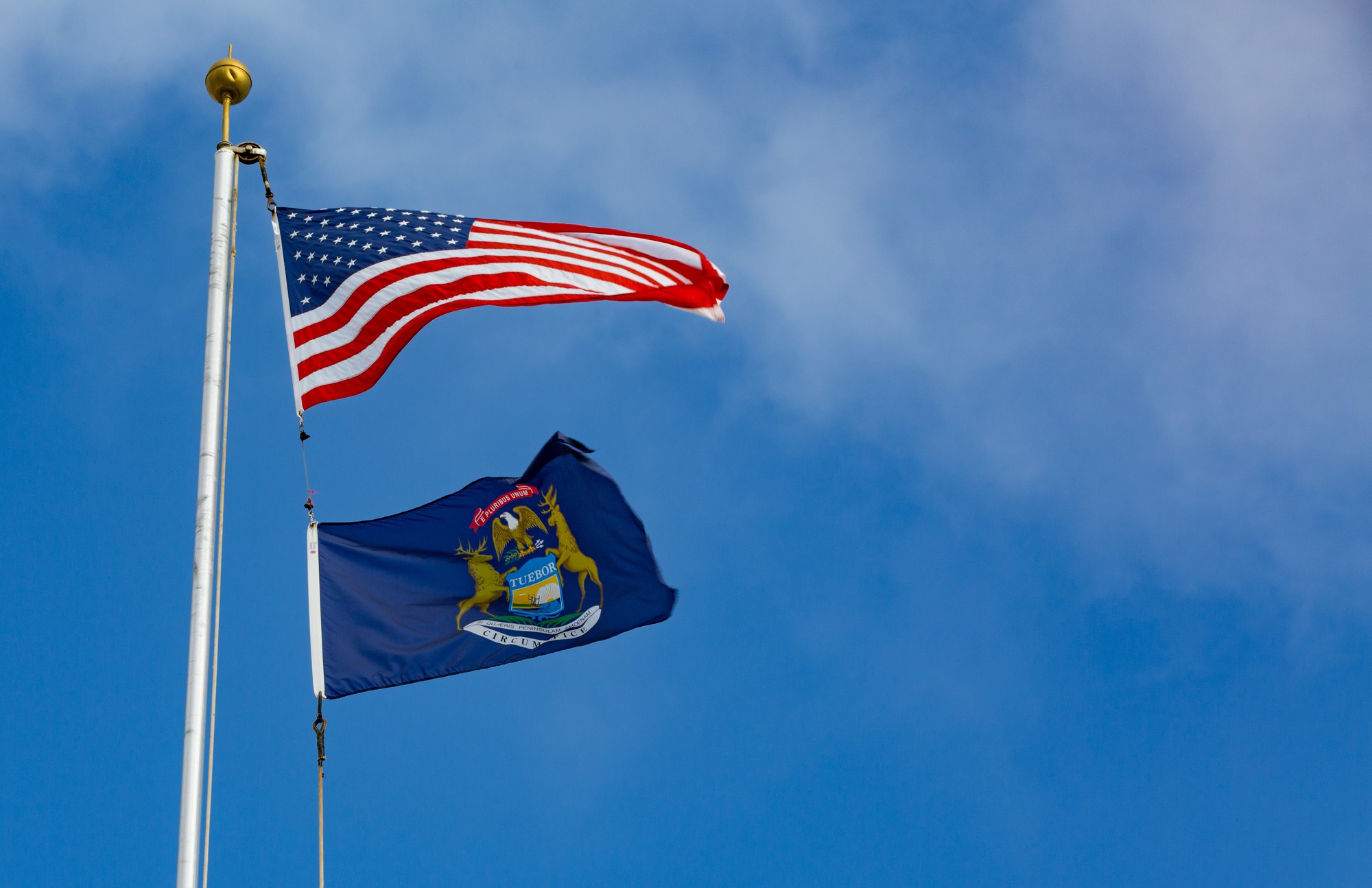 The American flag flying above the Michigan state flag against a blue sky