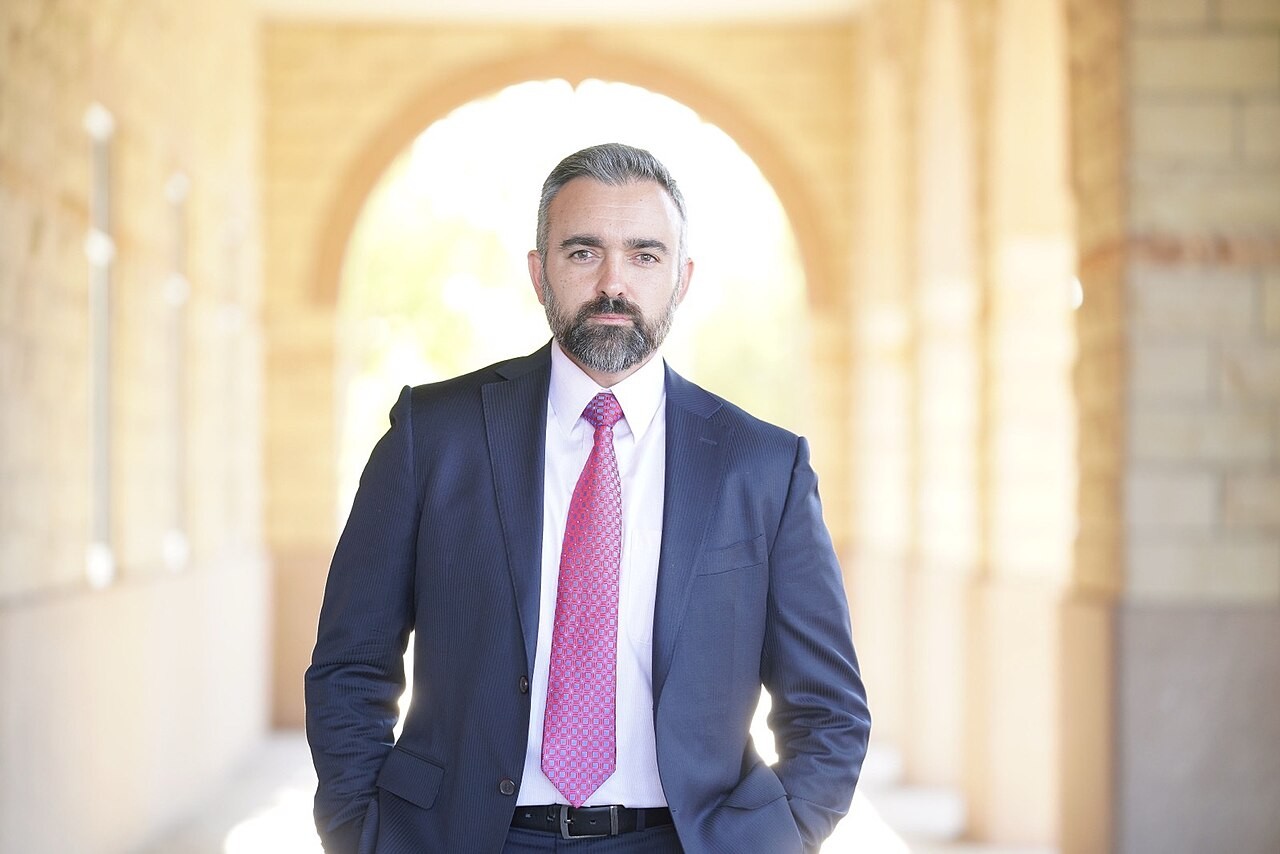 Portrait of New Mexico Attorney General Raúl Torrez in a dark suit and pink tie standing in an archway