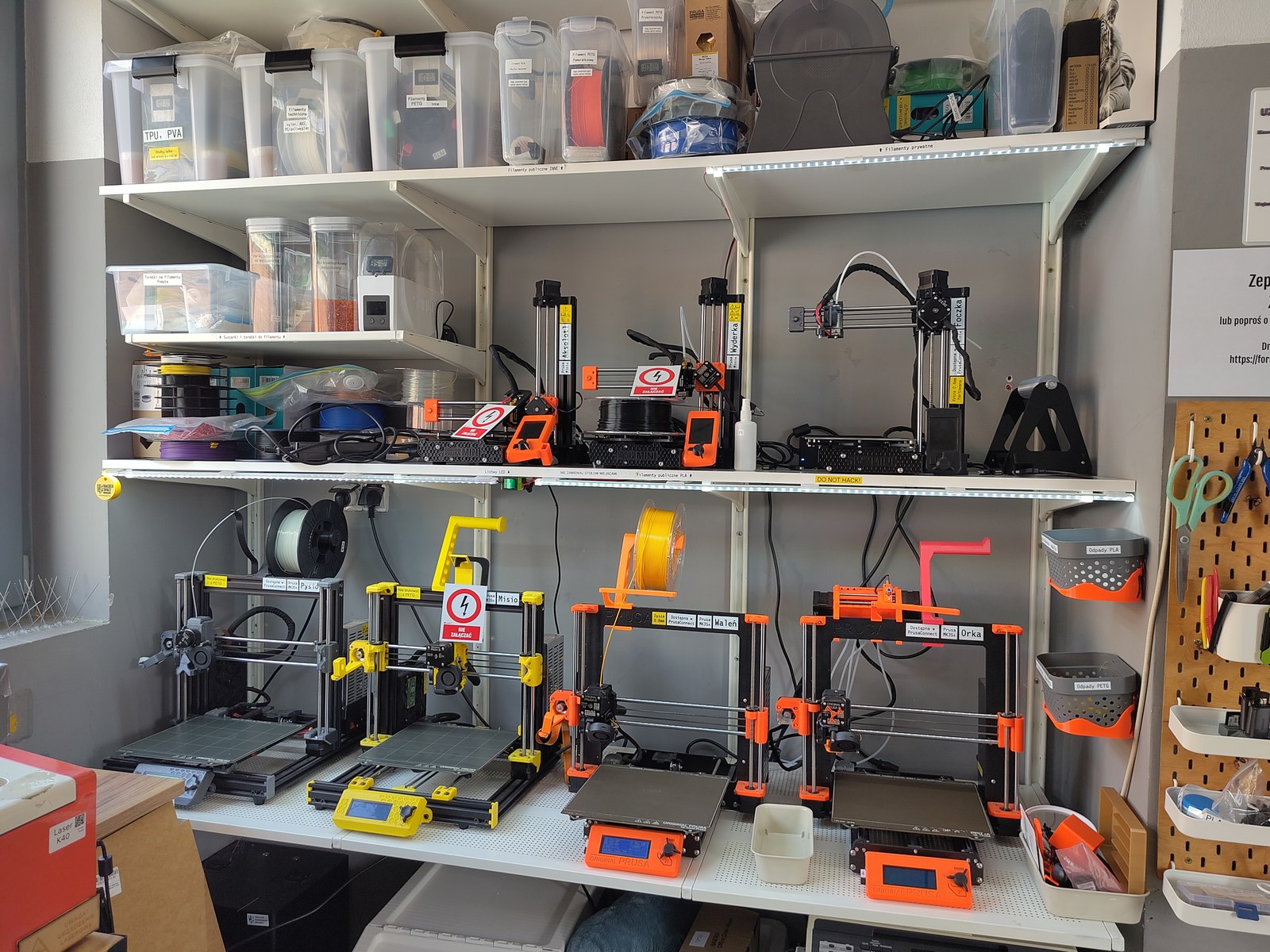 A shelf-mounted 3D printer farm of seven Prusa machines with filament spools and storage bins at Hackerspace Wroclaw