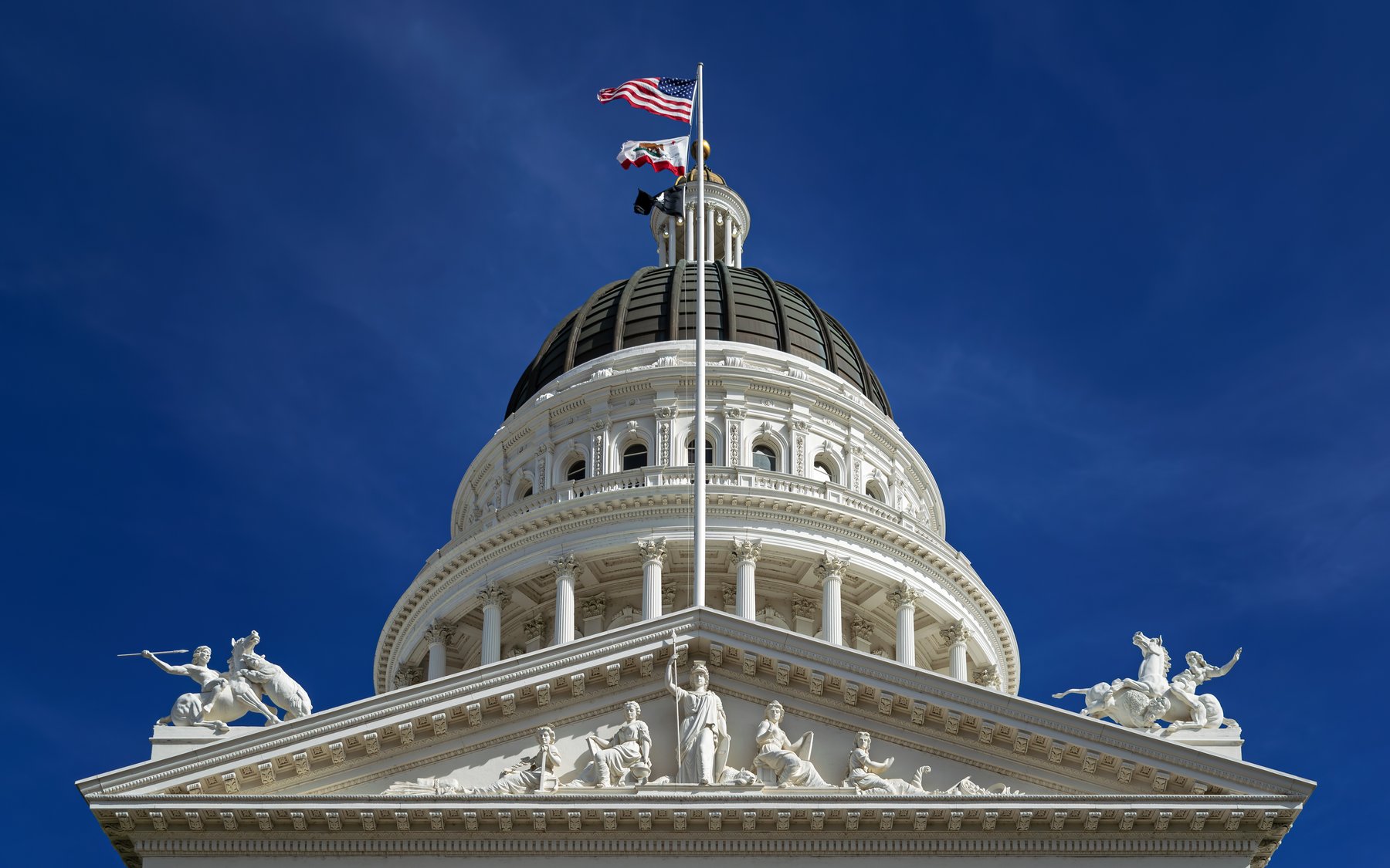The dome and pediment of the California State Capitol in Sacramento under a blue sky, with the U.S. and California flags flying