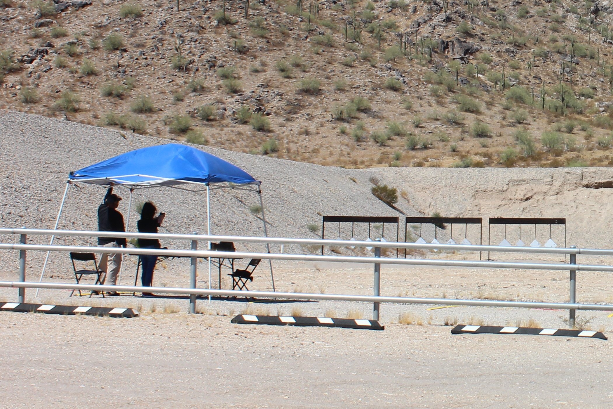 Two people at a covered shooting station at an outdoor pistol range with steel targets in the desert