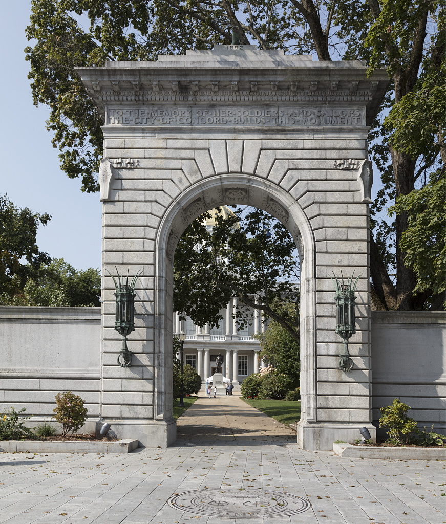 Stone memorial arch on the grounds of the New Hampshire State House in Concord, with the State House visible through the arch