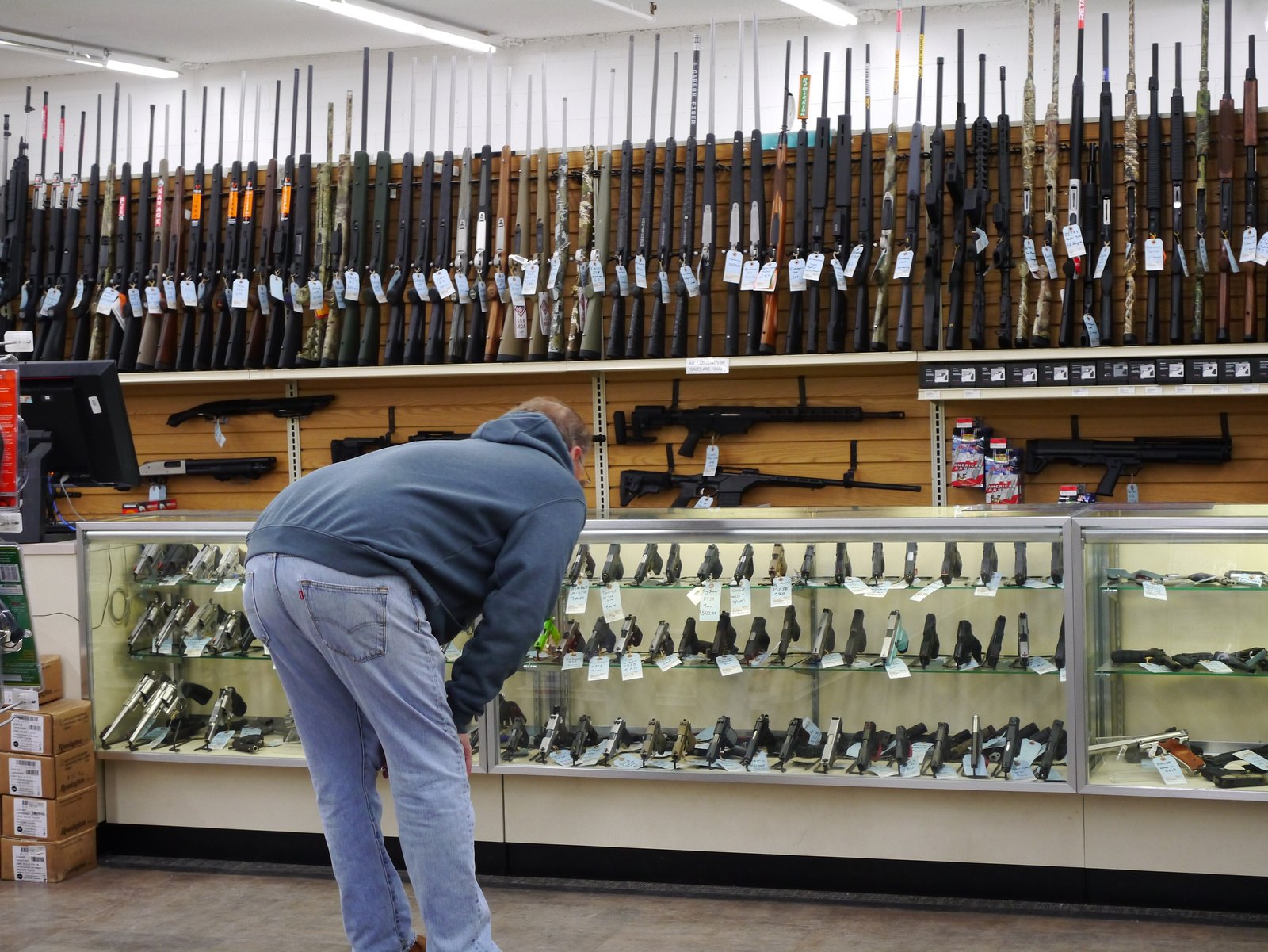 Customer looking at handguns in a glass display case beneath a wall of rifles in a retail store