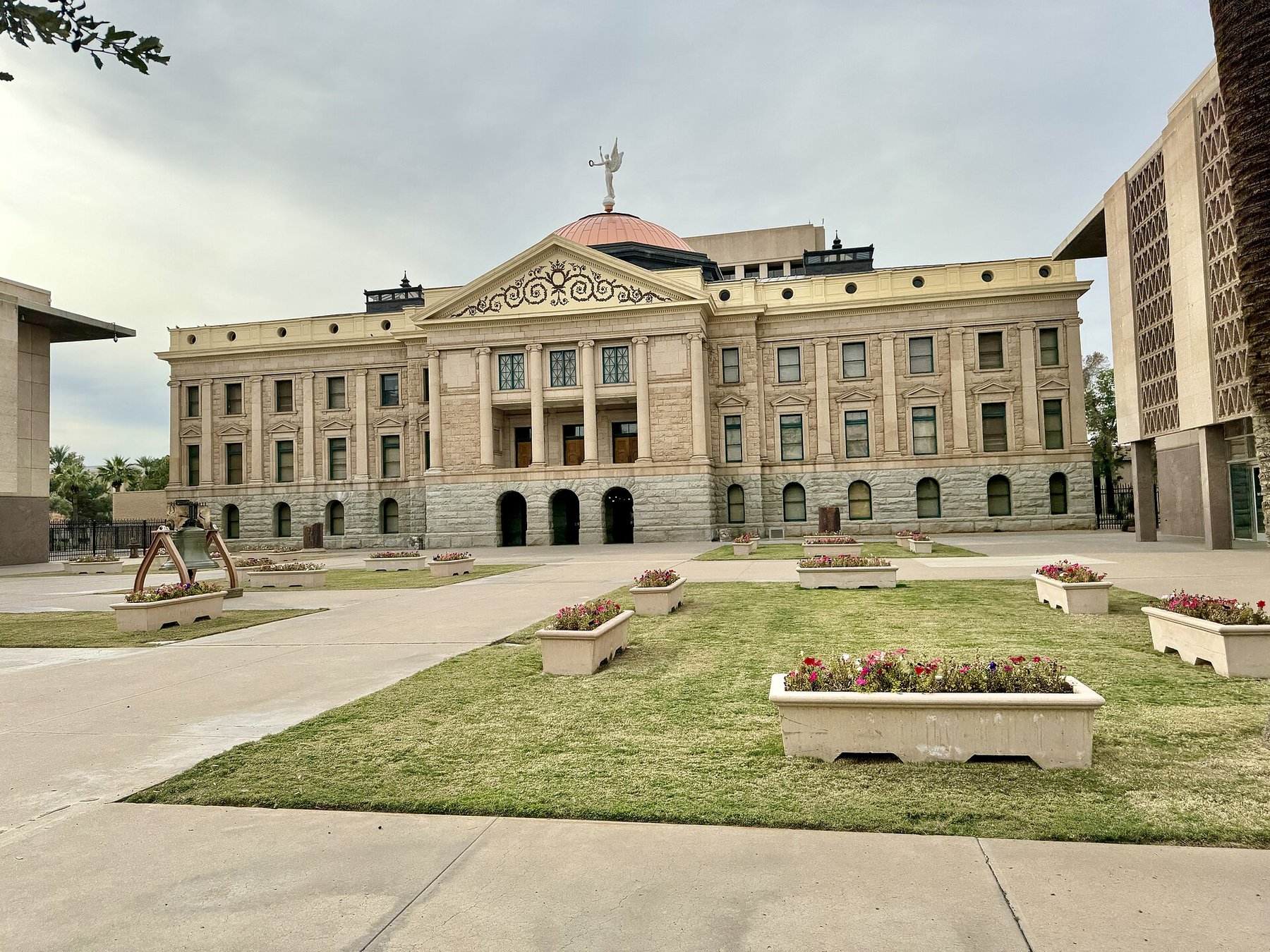 The historic Arizona State Capitol building in Phoenix with its copper dome and winged figure, seen across a lawn with flower planters