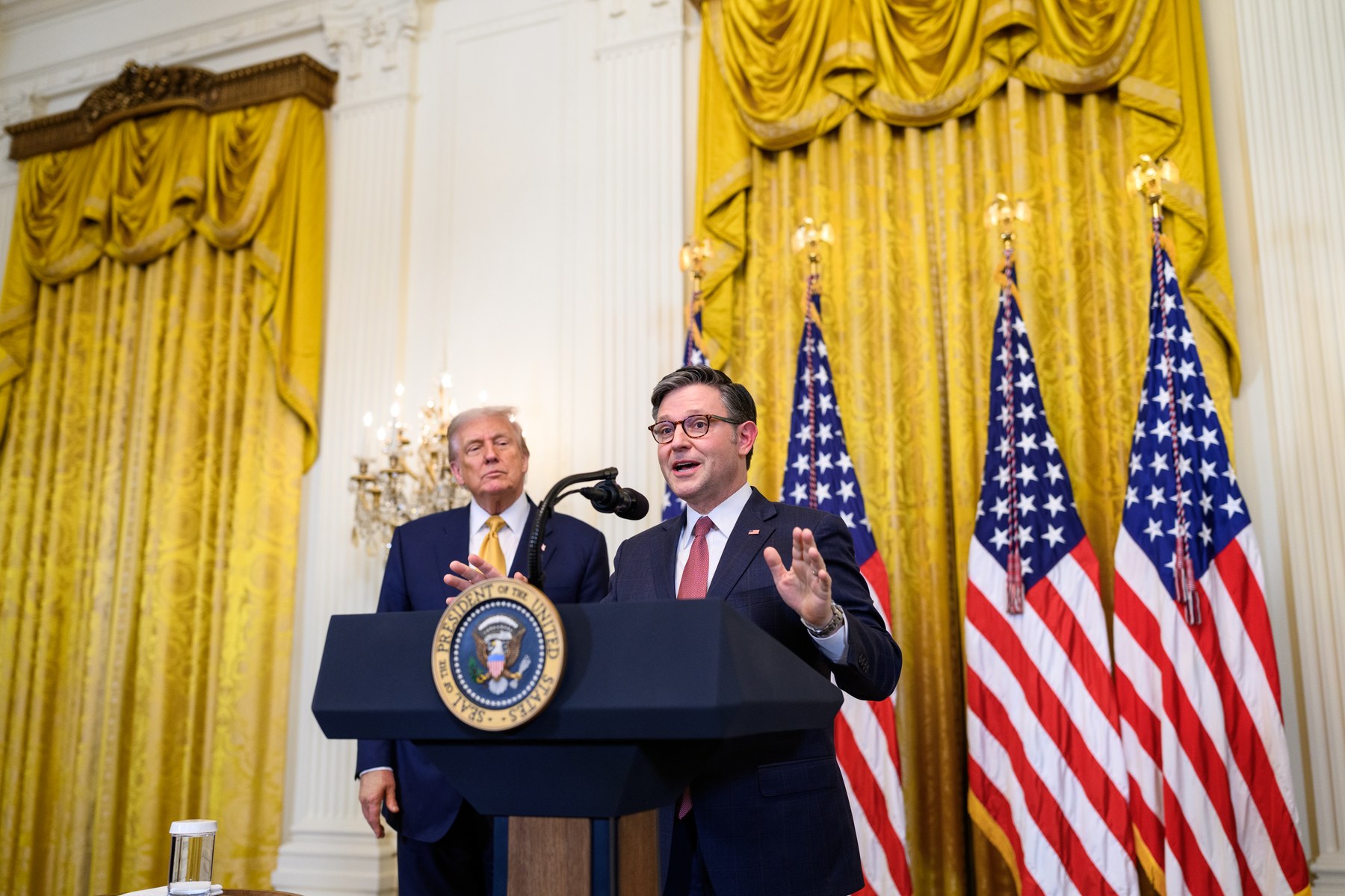 Speaker of the House Mike Johnson speaking at a podium with President Donald Trump standing behind him at the White House