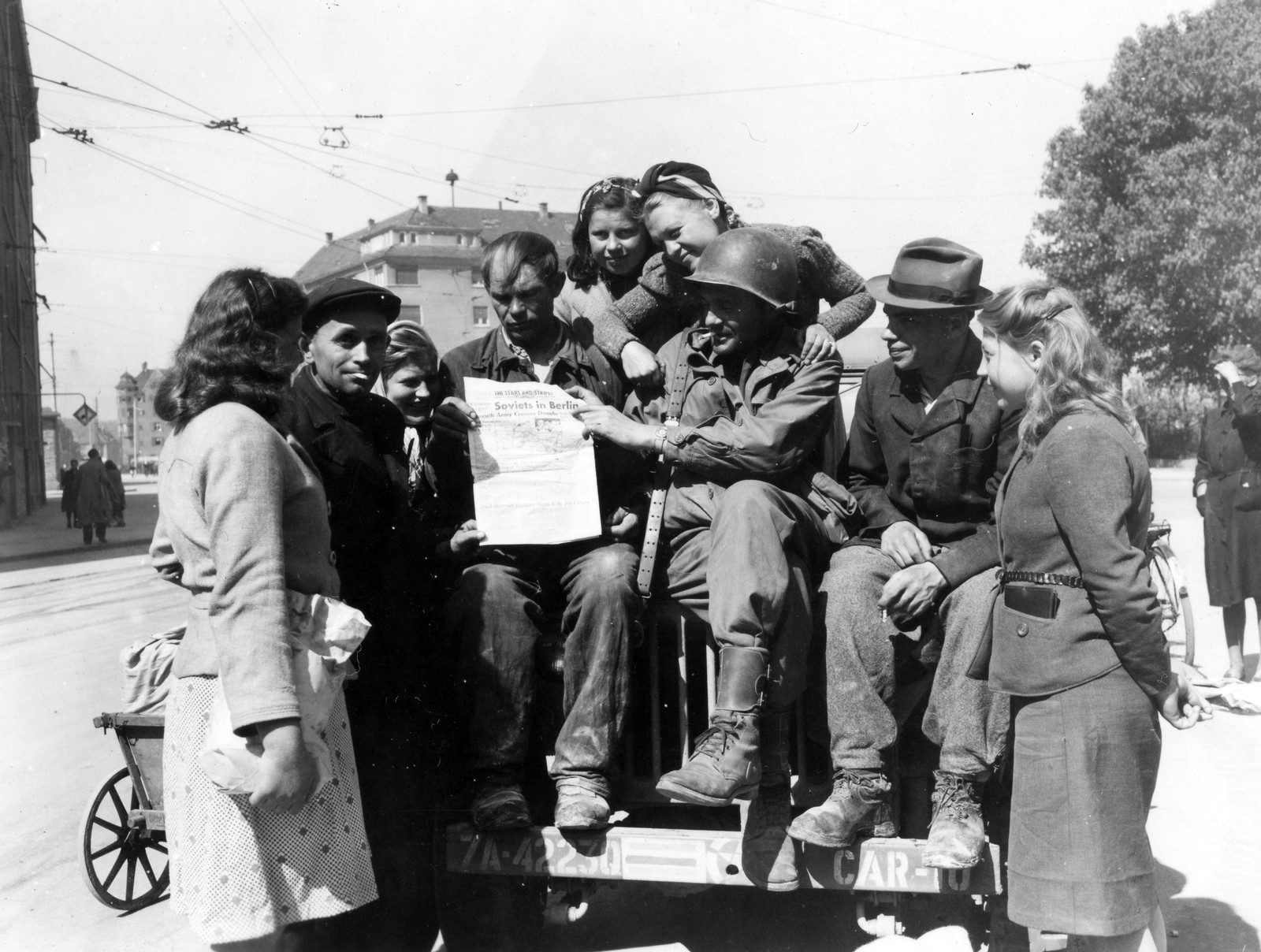 An American soldier sitting on the tailgate of a truck in Ulm, Germany, in April 1945, showing a copy of Stars and Stripes with the headline Soviets in Berlin to a group of newly liberated civilians
