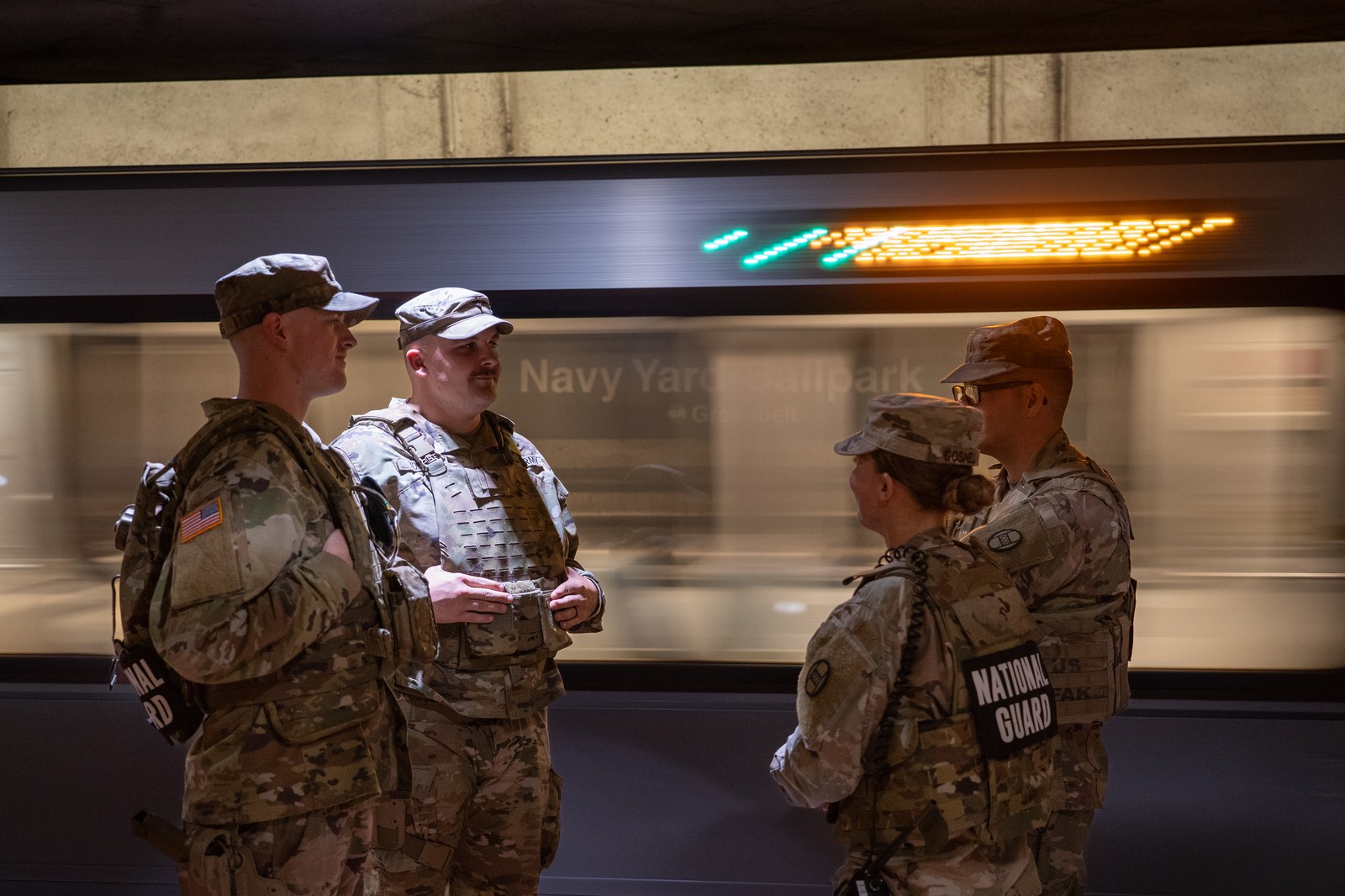 Four National Guard soldiers in camouflage uniforms standing on the platform of the Navy Yard Ballpark Metro station in Washington, D.C., as a train passes behind them