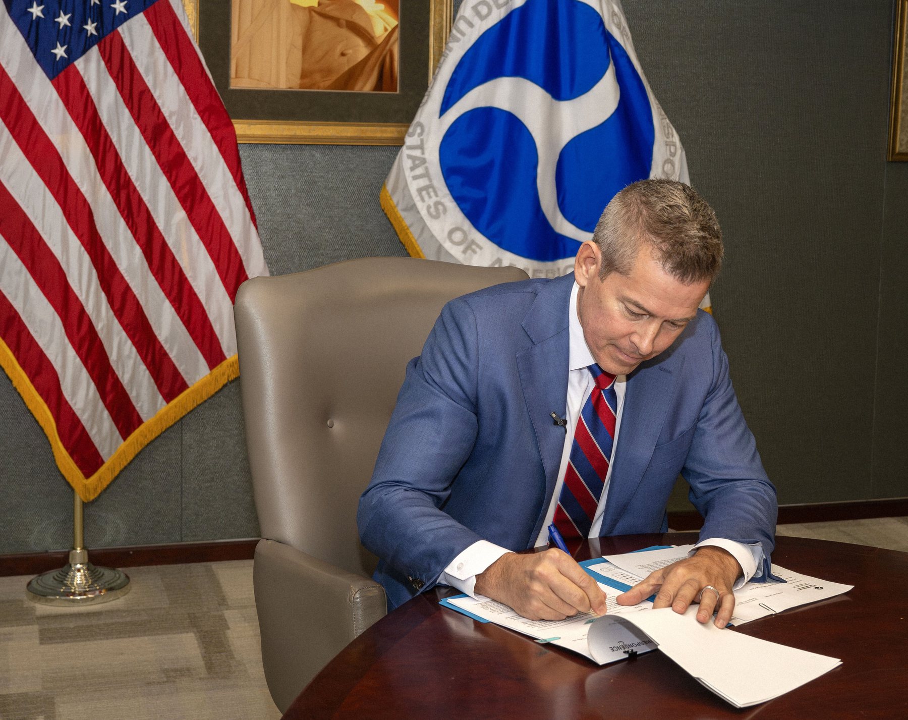 Transportation Secretary Sean Duffy seated at a desk signing a memorandum on fuel economy standards, with an American flag and the DOT flag behind him