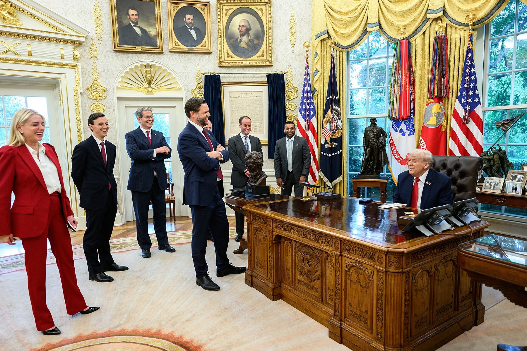 President Donald Trump seated at the Resolute Desk in the Oval Office with Vice President JD Vance and aides standing nearby