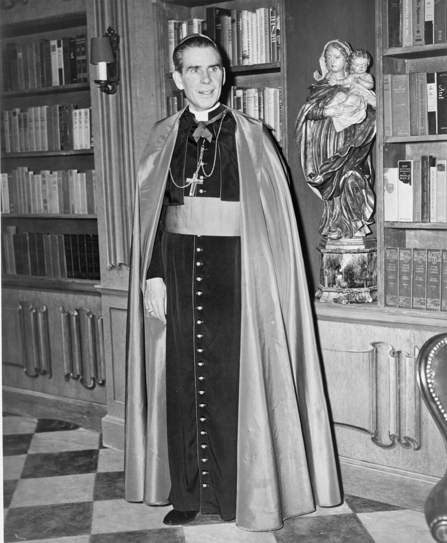 Bishop Fulton J. Sheen in cassock and cape standing on the library set of his television program in 1952