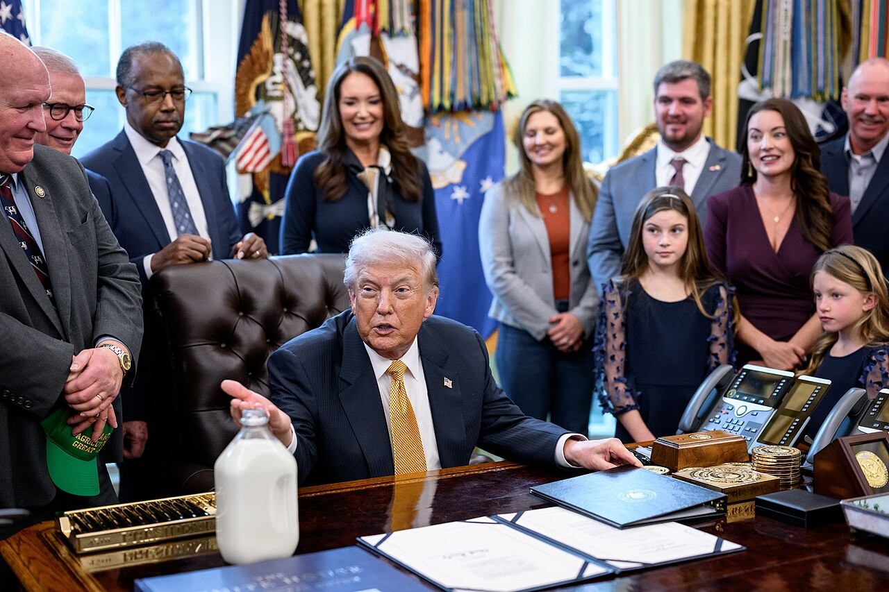 President Trump speaking at the Resolute Desk with dairy farmers, Agriculture Secretary Rollins and HHS Secretary Kennedy standing behind him