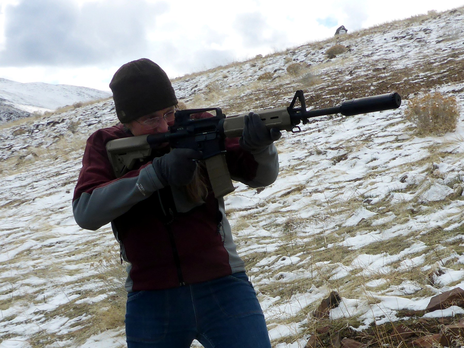 A shooter in a knit cap and shooting glasses aiming a suppressed AR-15 rifle on a snowy hillside