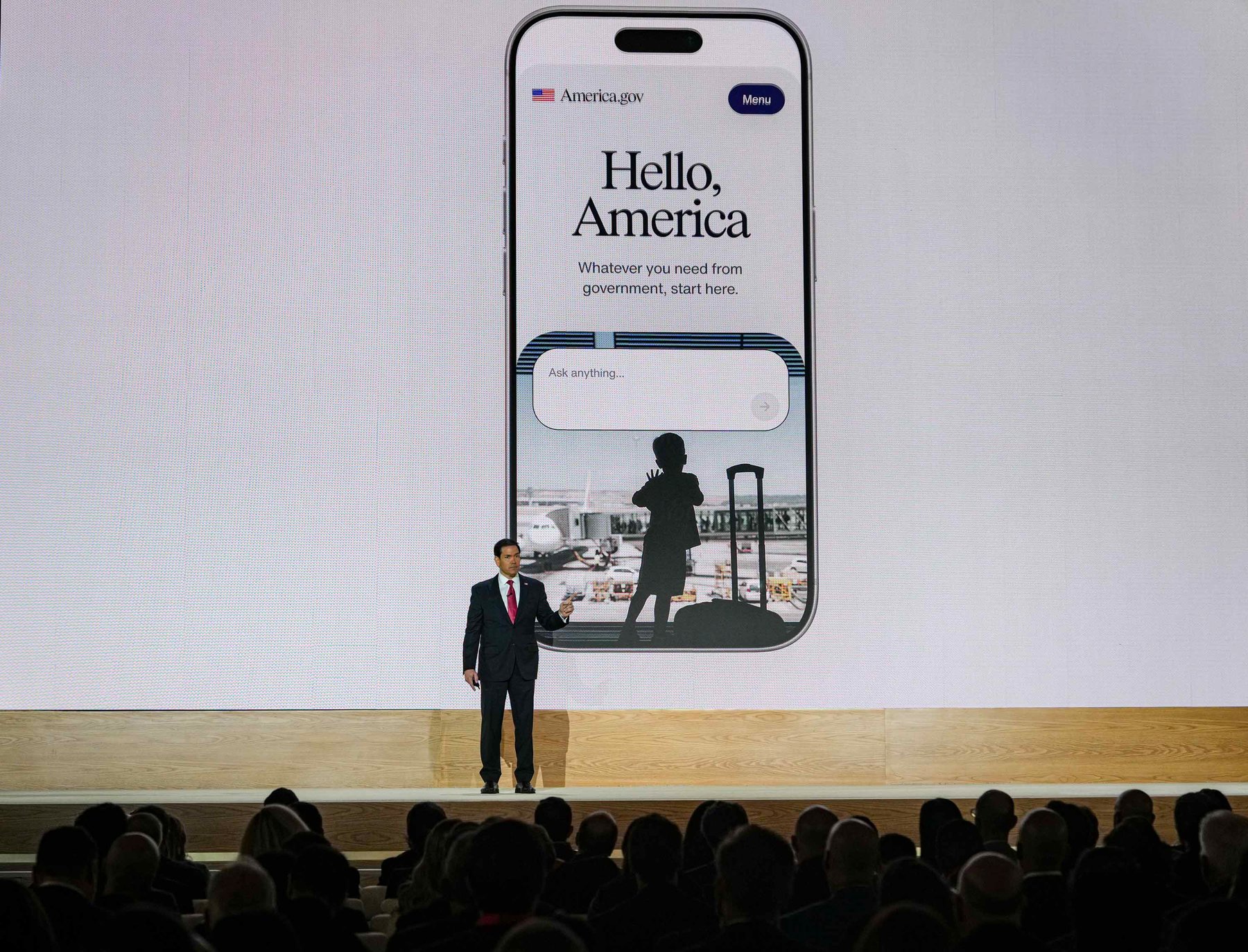 Secretary of State Marco Rubio on stage in front of a giant screen showing the America.gov app with the words Hello, America and an Ask anything search box