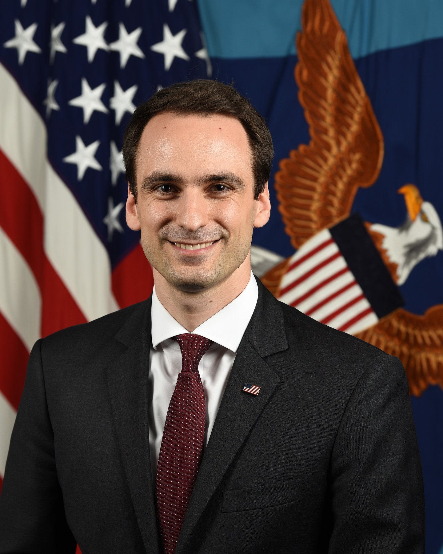 Official portrait of Michael Kratsios in a dark suit and red tie in front of an American flag