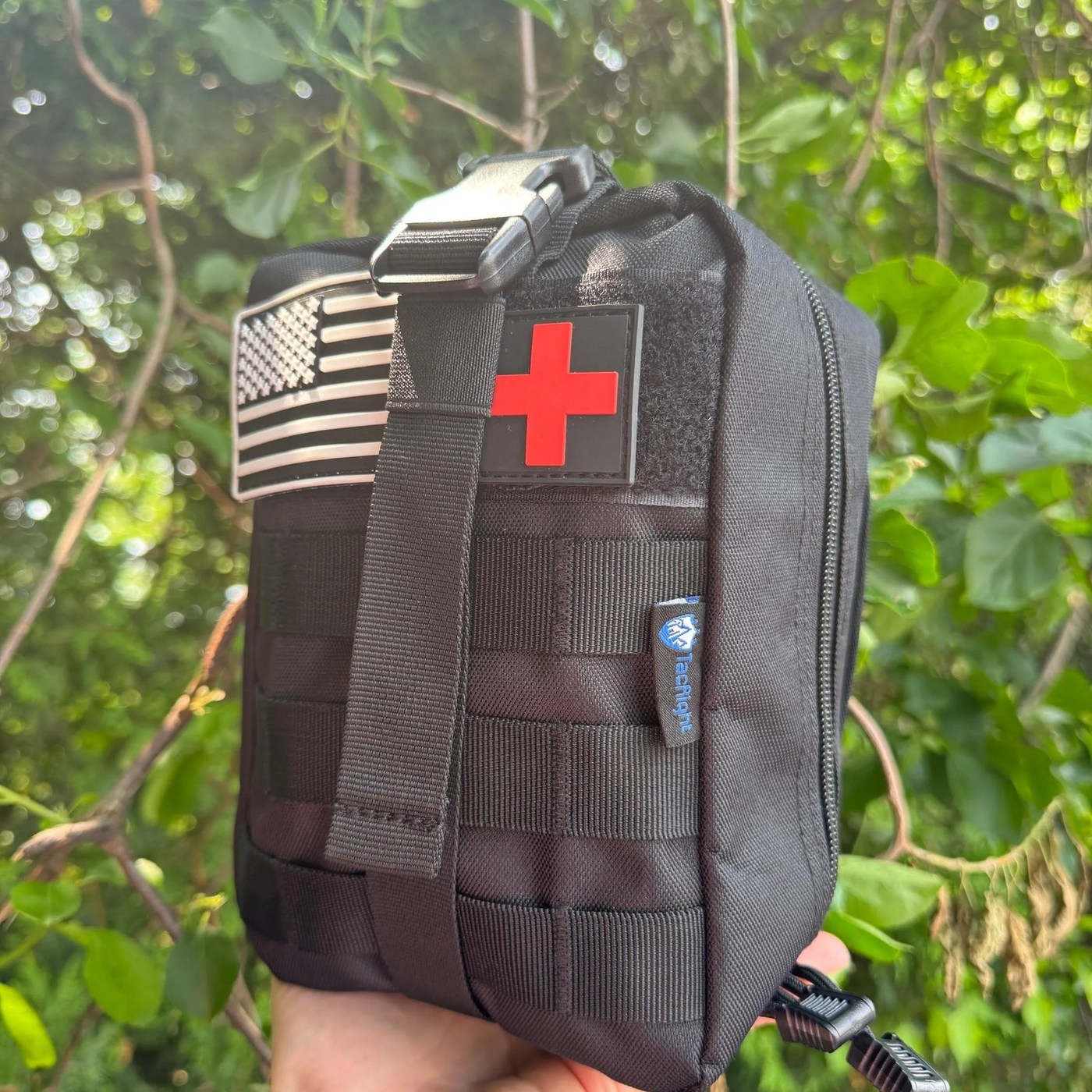 The Trauma First Aid Kit held in one hand, showing the MOLLE webbing and patches on the front