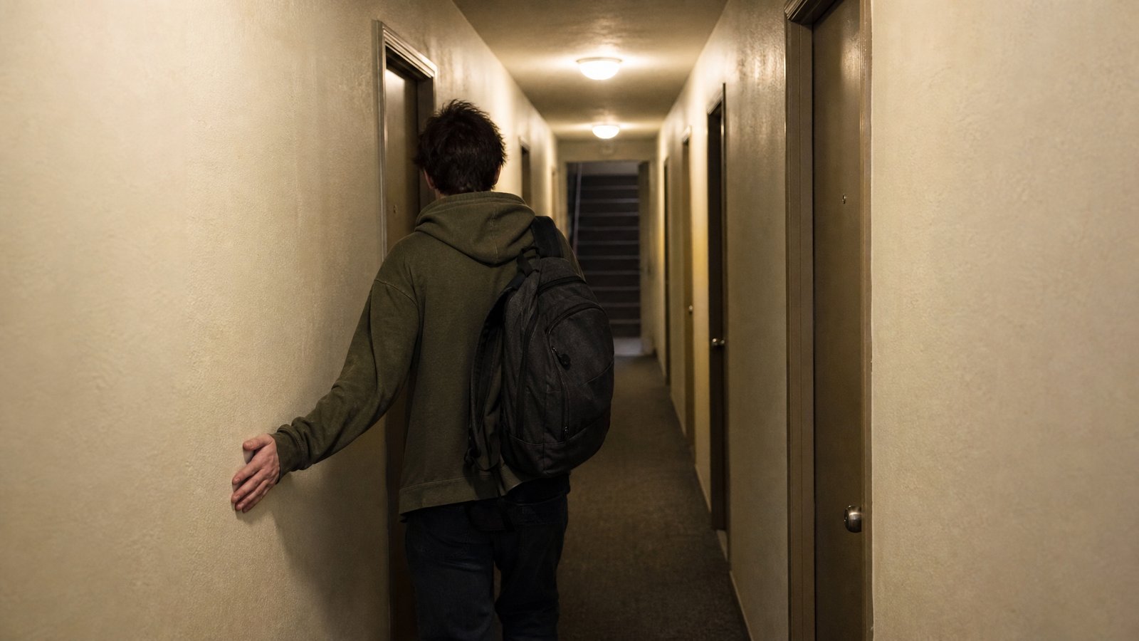A person walking down an apartment hallway toward a stairwell door, one hand trailing along the wall to count doors