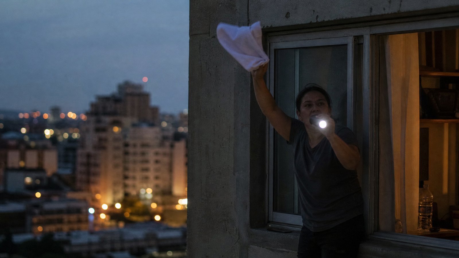 A person at an apartment window at dusk holding a lit flashlight and waving a white cloth