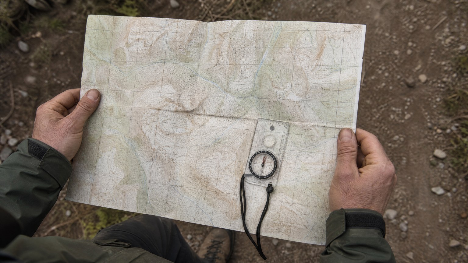 Overhead view of weathered hands holding an open folded paper topographic map flat with a small clear-baseplate compass resting on top of it and a dirt trail visible at the edge of the frame