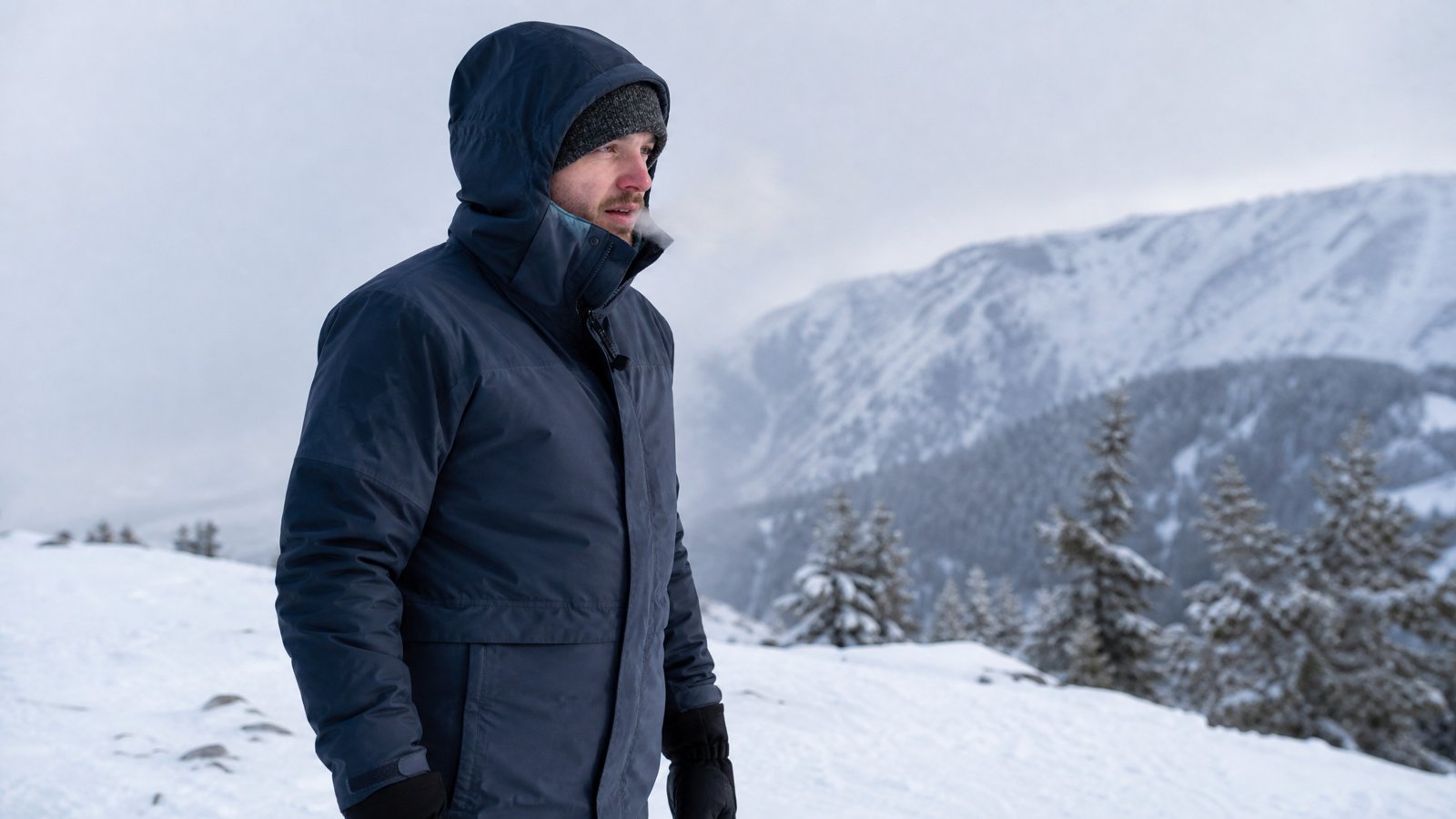 A person in a hooded shell jacket, knit hat and gloves standing on a snow-covered ridge with breath visible in freezing air
