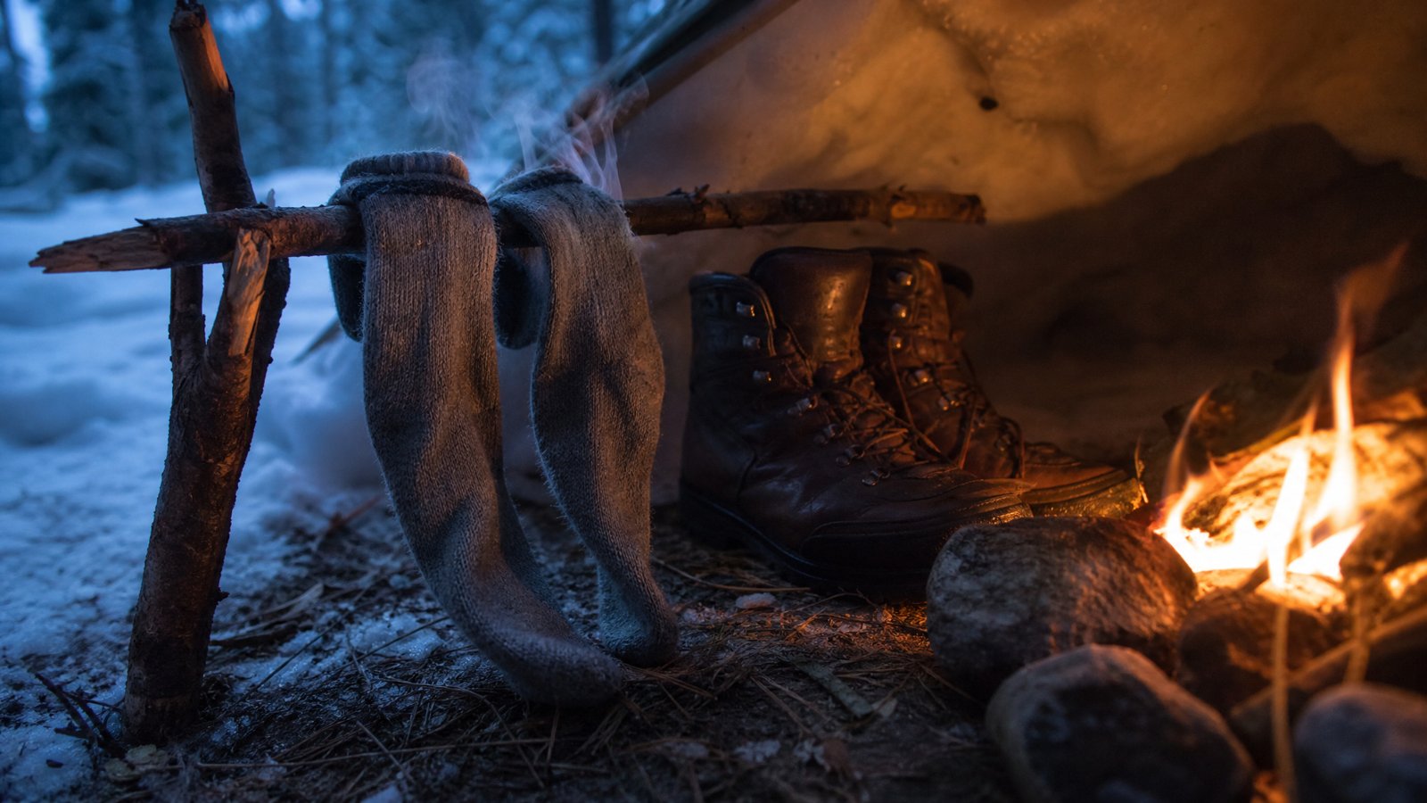 Wet wool socks hung on a stick to dry beside a small campfire at dusk with damp leather boots warming nearby in the snow